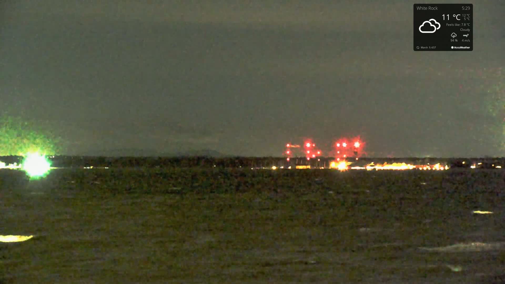 A nighttime view of a body of water with a distant shoreline showing lights and what appear to be red flares under a cloudy sky.