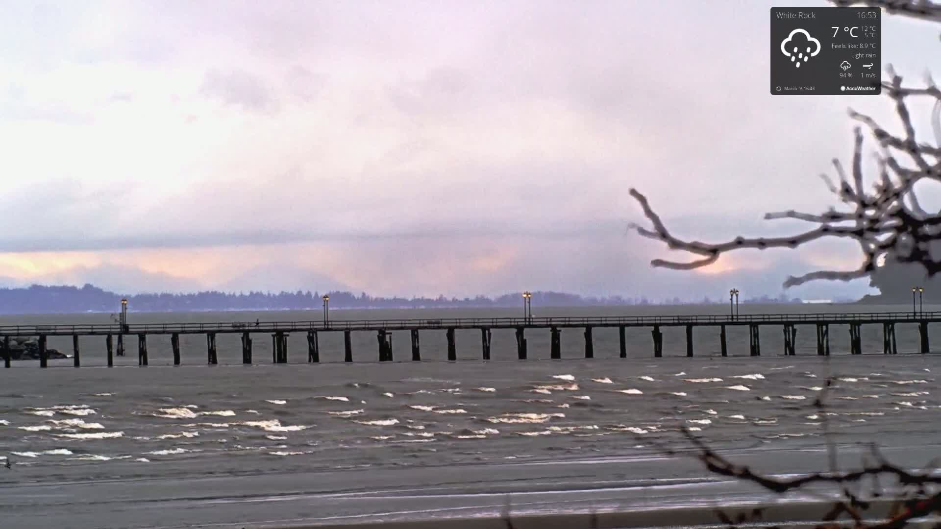 A long pier extends over a choppy, gray ocean under a cloudy, light-pink sky during a light rain.
