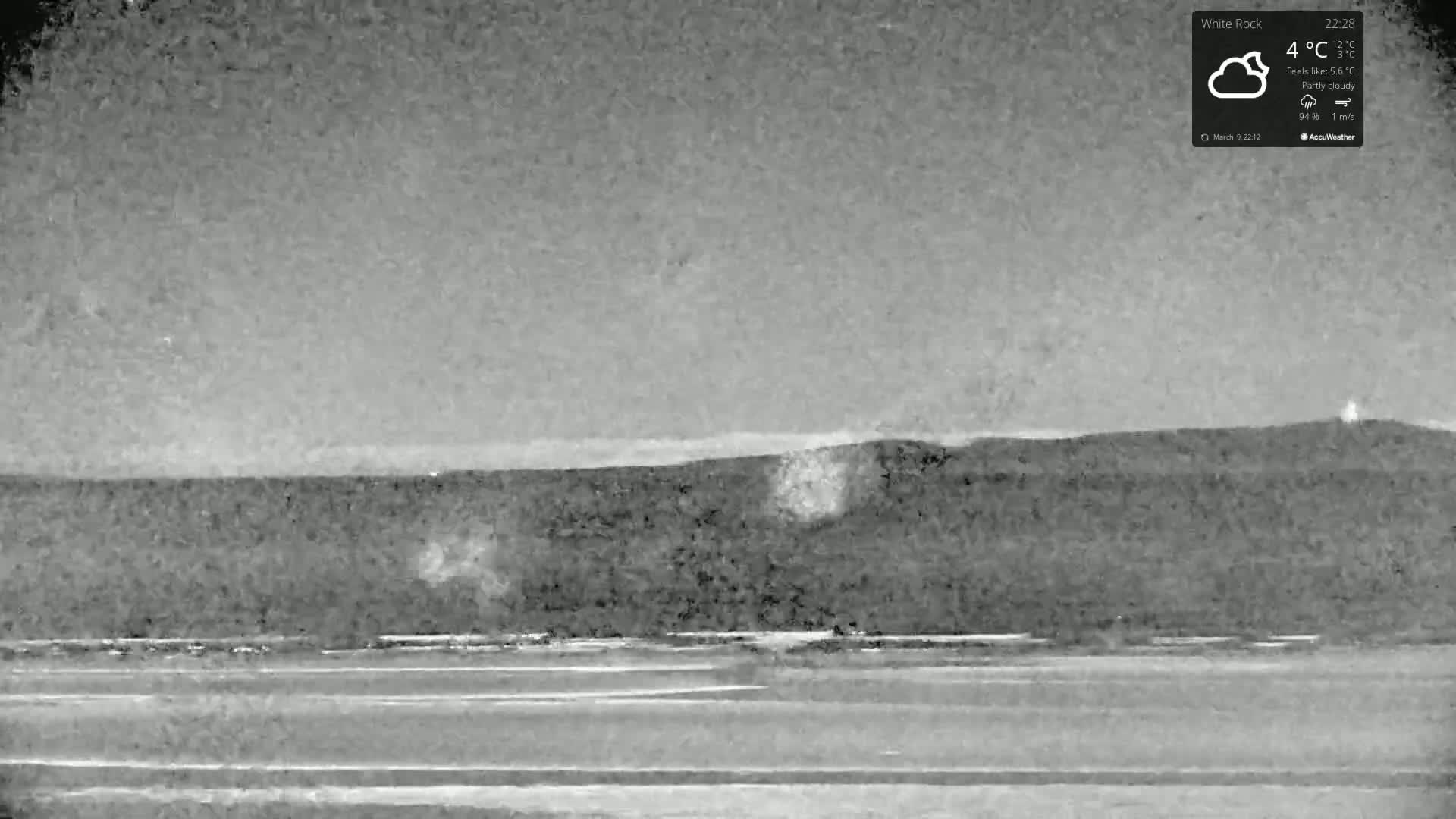 A nighttime, partly cloudy scene shows a flat expanse of land leading to a dark hill under a light gray sky.