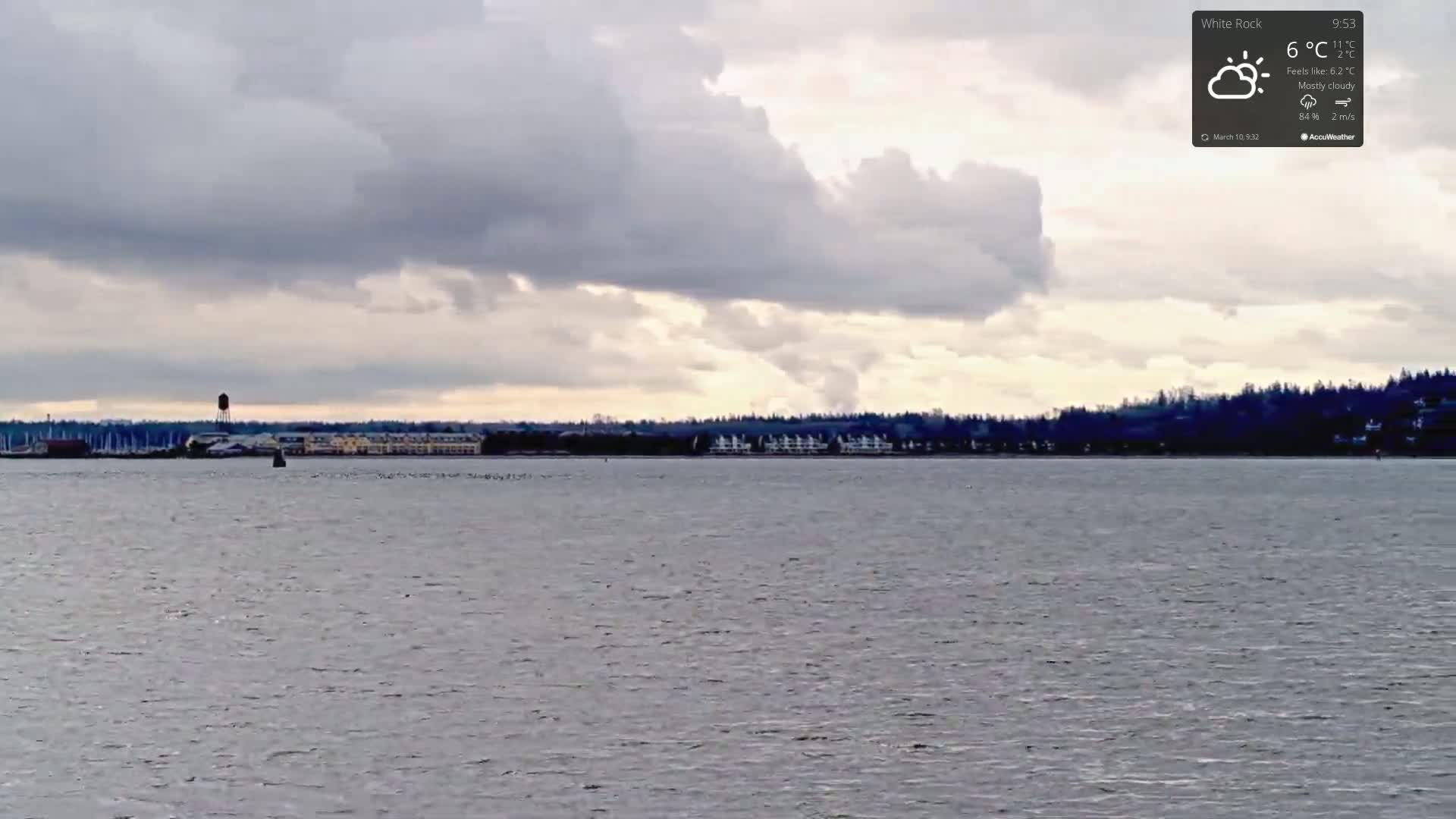 A mostly cloudy sky hangs over a calm body of water, with a distant shoreline featuring buildings and trees.