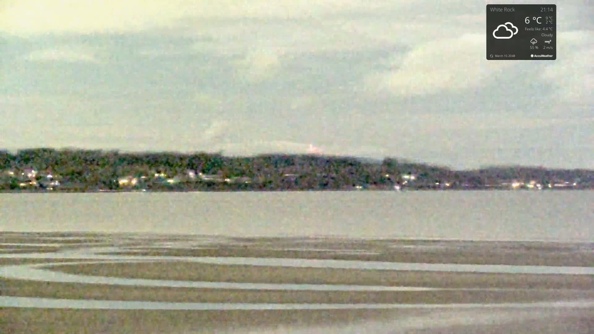 A calm body of water, possibly a lake or bay, with a gently sloping, patterned shoreline in the foreground, overlooks a dark, tree-covered landmass under a cloudy sky.