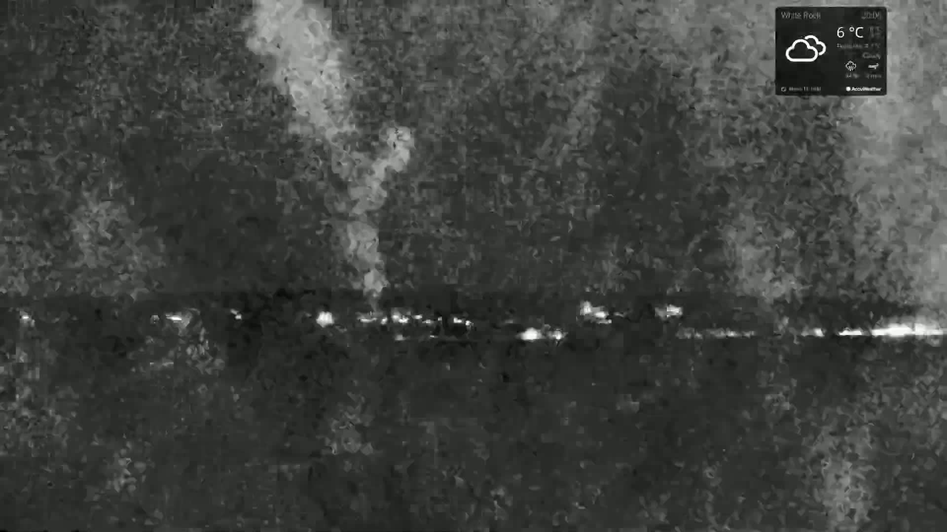 A nighttime, cloudy scene shows a line of lights in the distance with a plume of smoke or fog rising in the center.