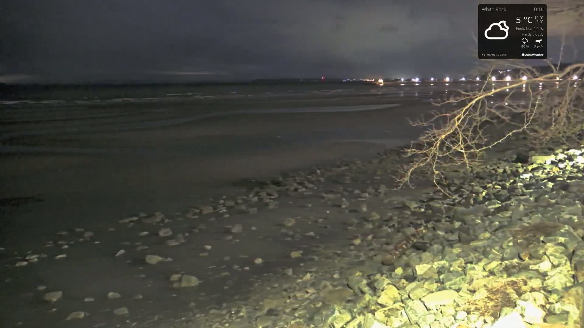 A dark, partly cloudy night scene shows a rocky shoreline and a calm, dark body of water under a mostly dark sky, with distant lights visible on the opposite shore.