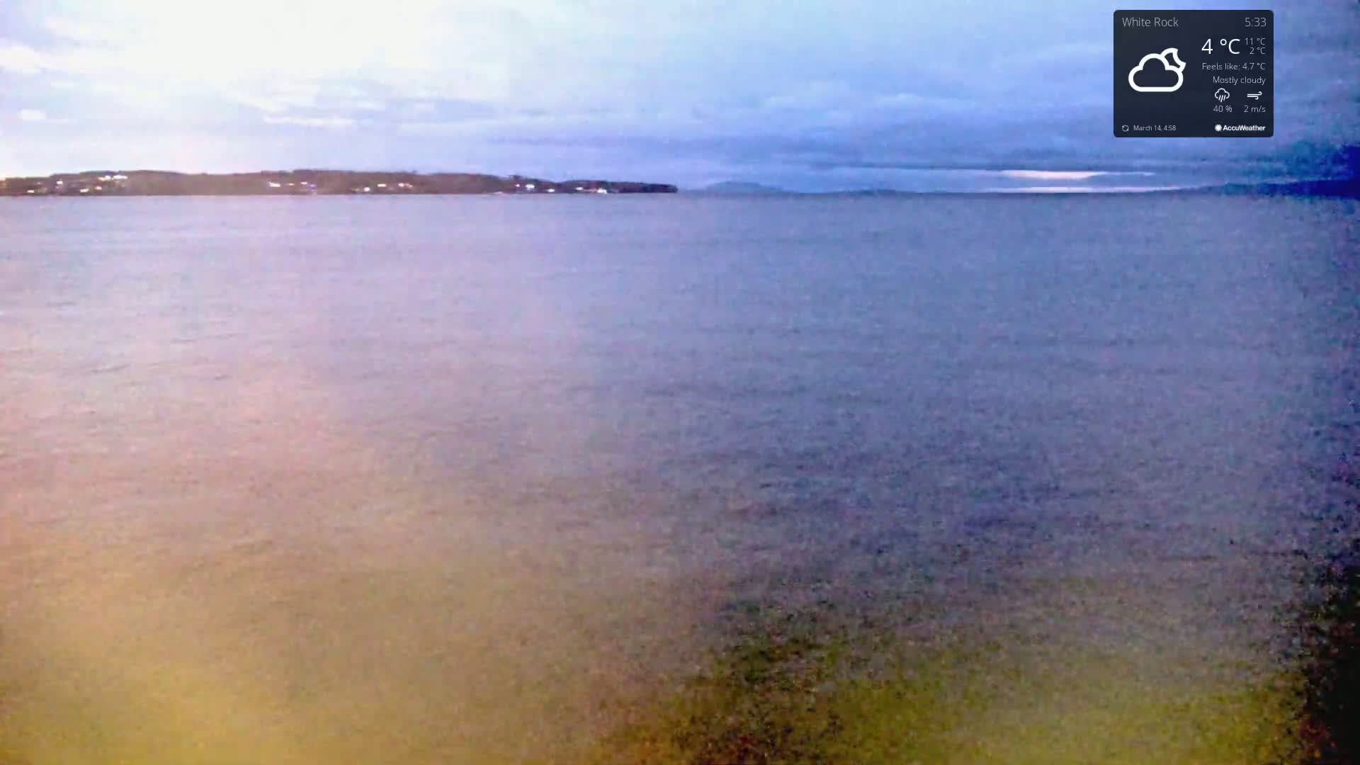 A mostly cloudy, cool day is shown overlooking a calm body of water with a distant shoreline.