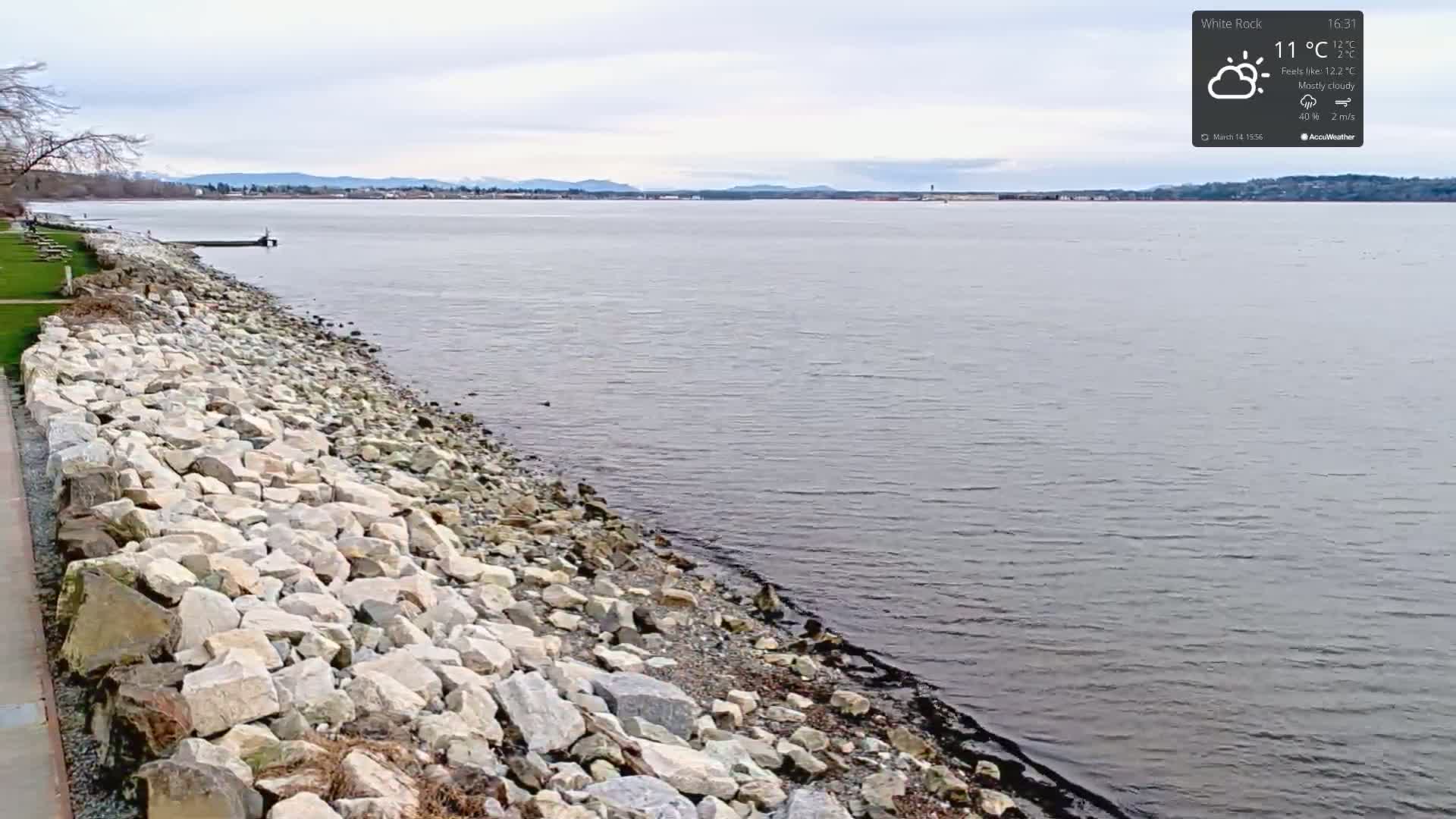 A rocky shoreline stretches along a calm body of water under a mostly cloudy sky.