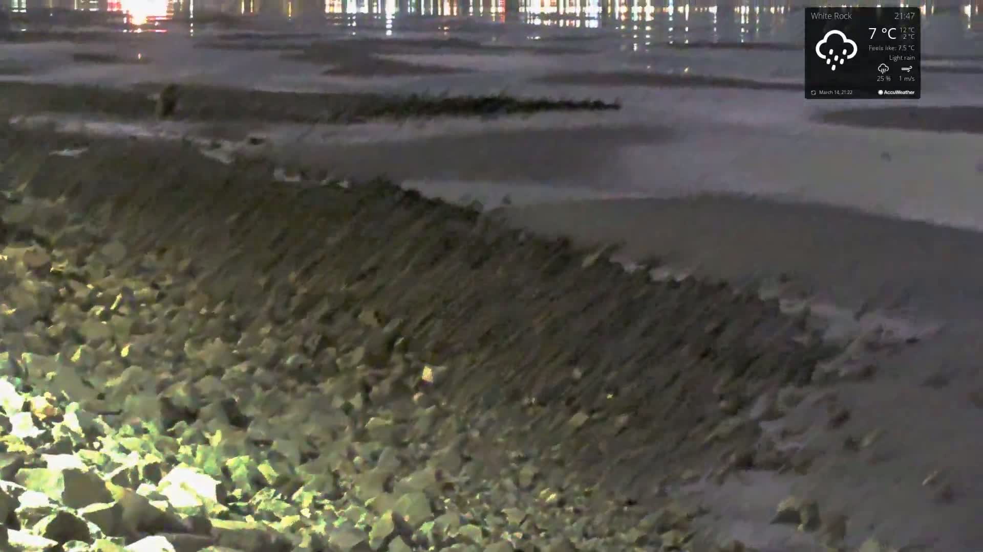 A rocky shoreline at night under light rain, with city lights visible in the distance across the water.