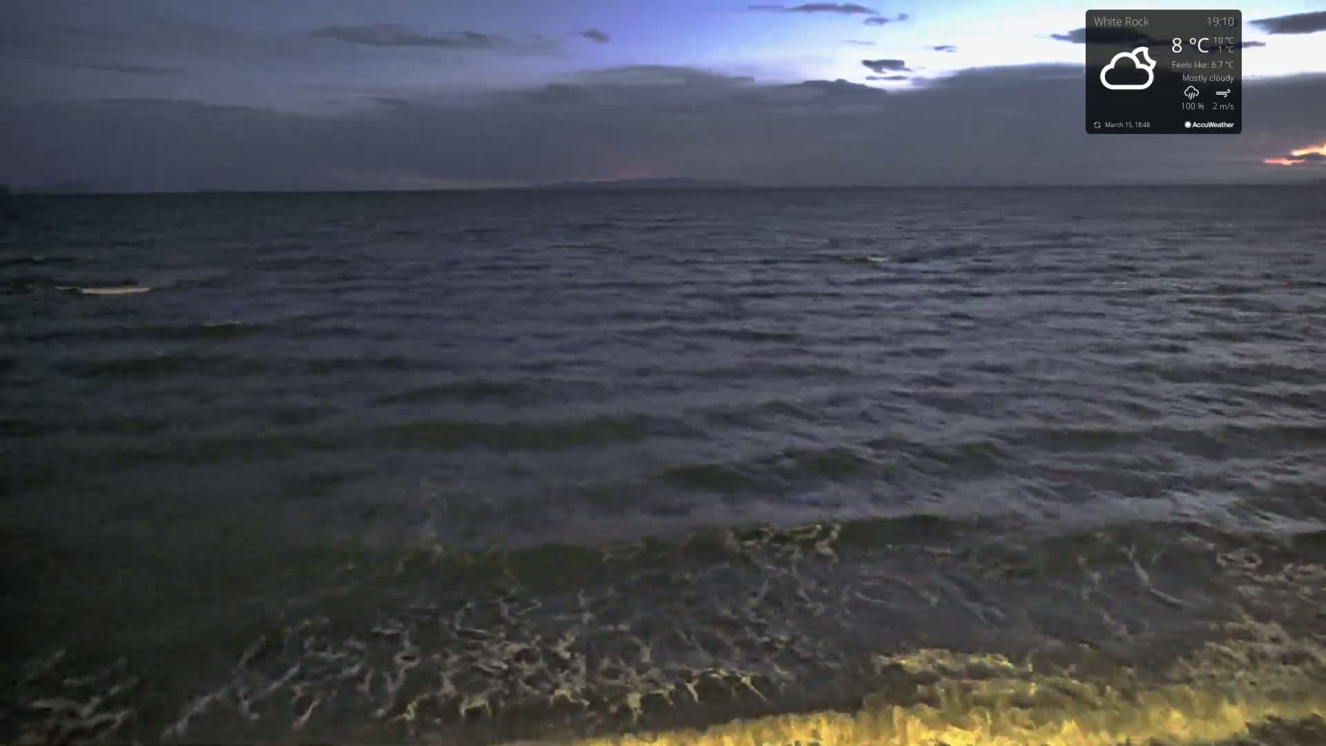 A dark, mostly cloudy evening shows gentle waves lapping at a shoreline under a dusky sky.