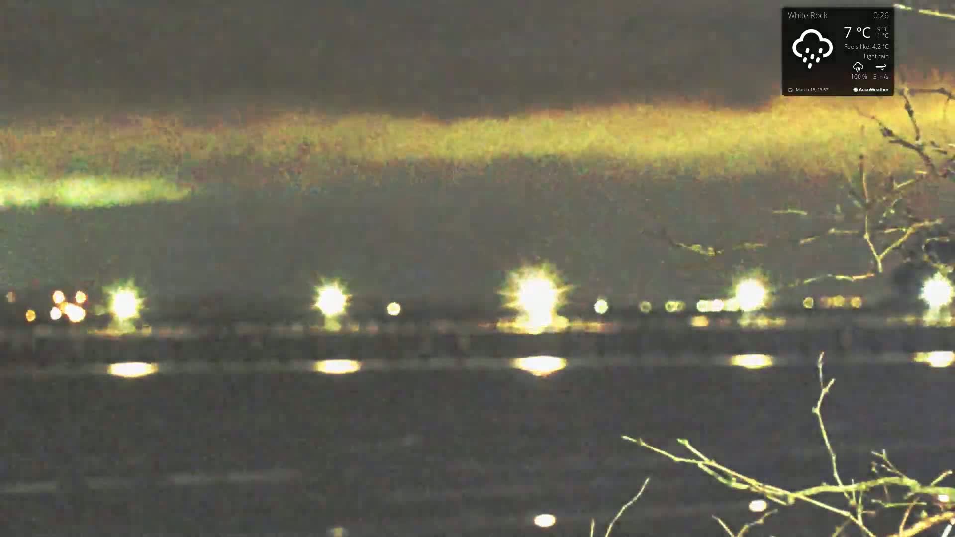 A nighttime view of a body of water with distant lights reflecting on the surface, under a dark sky with light rain.