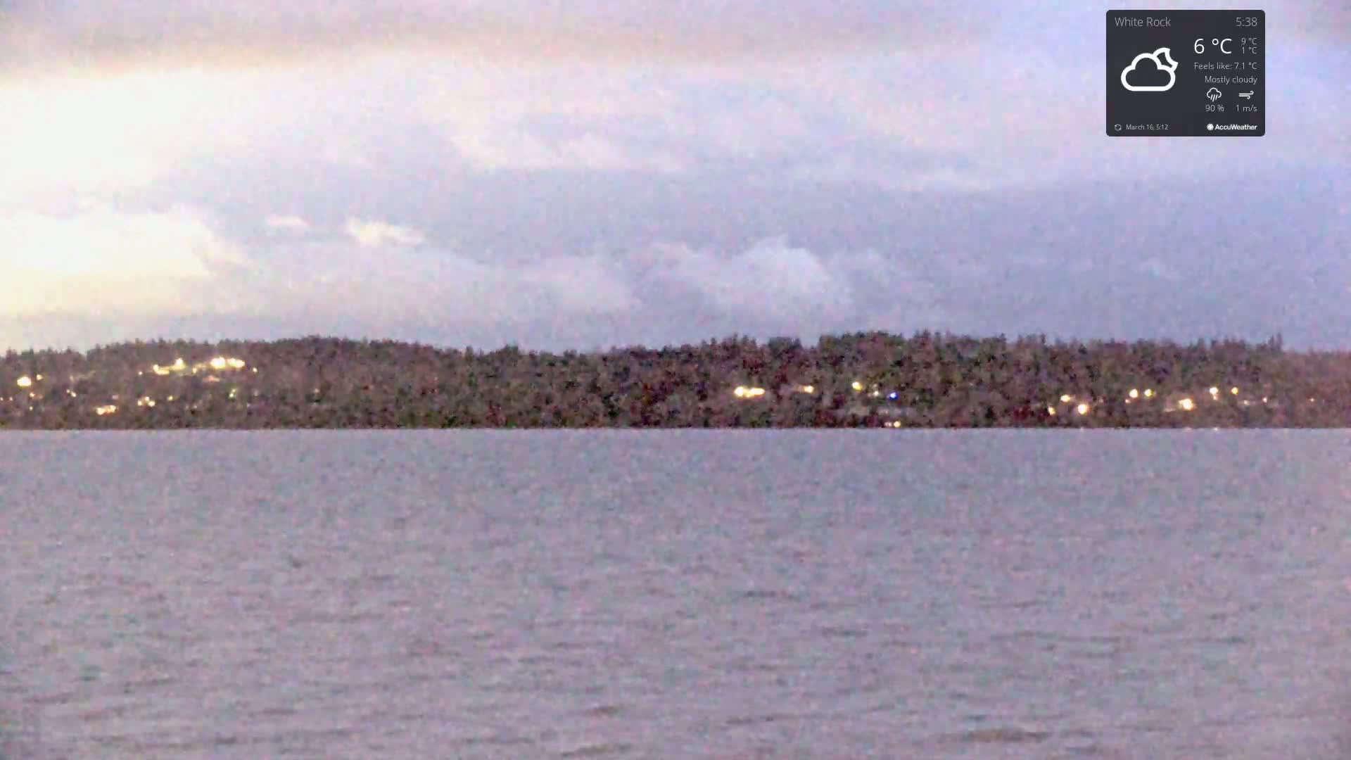 A calm body of water stretches to a distant shoreline of dark trees speckled with lights under a mostly cloudy, dusky sky.