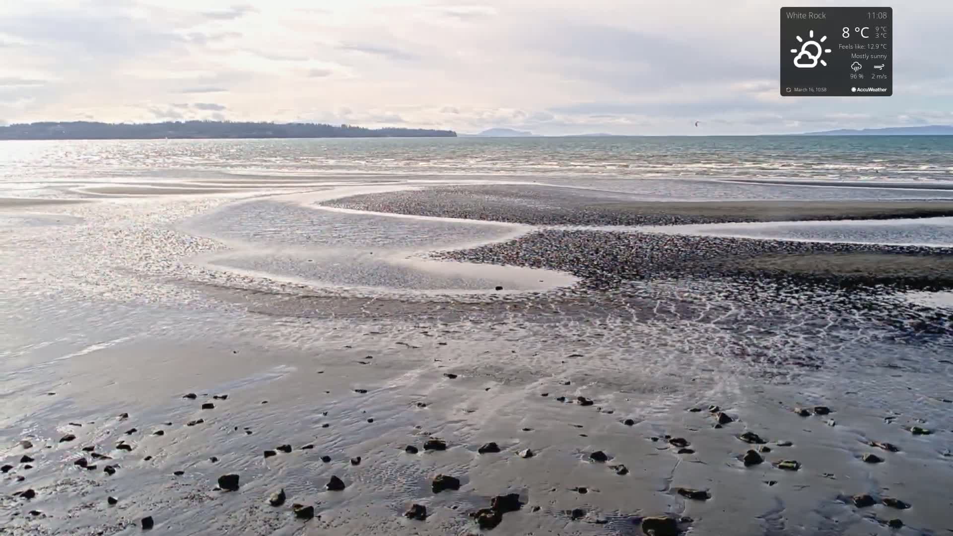A mostly sunny day reveals a gently sloping beach with small, dark rocks and shallow, meandering channels of water receding from the shore.