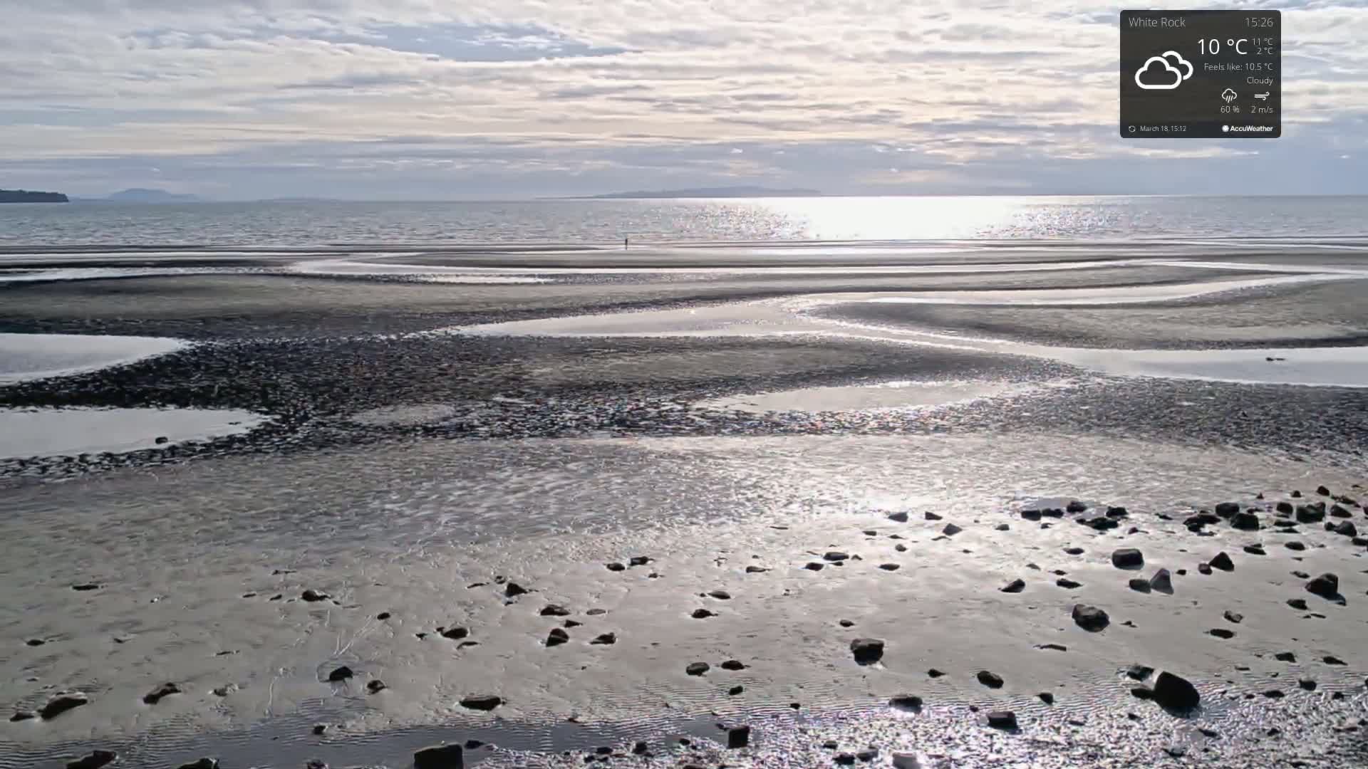 A mostly sunny, cloudy day reveals a wide expanse of tidal flats with scattered rocks and shallow pools of water reflecting sunlight, while a calm sea stretches to a distant shoreline.