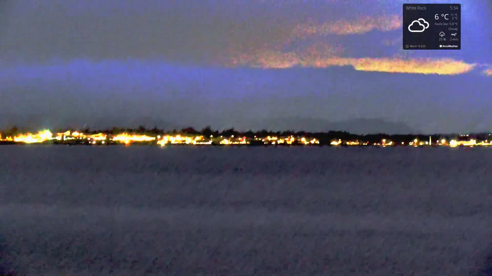 A nighttime view of a town's lights across a body of water under a cloudy sky.