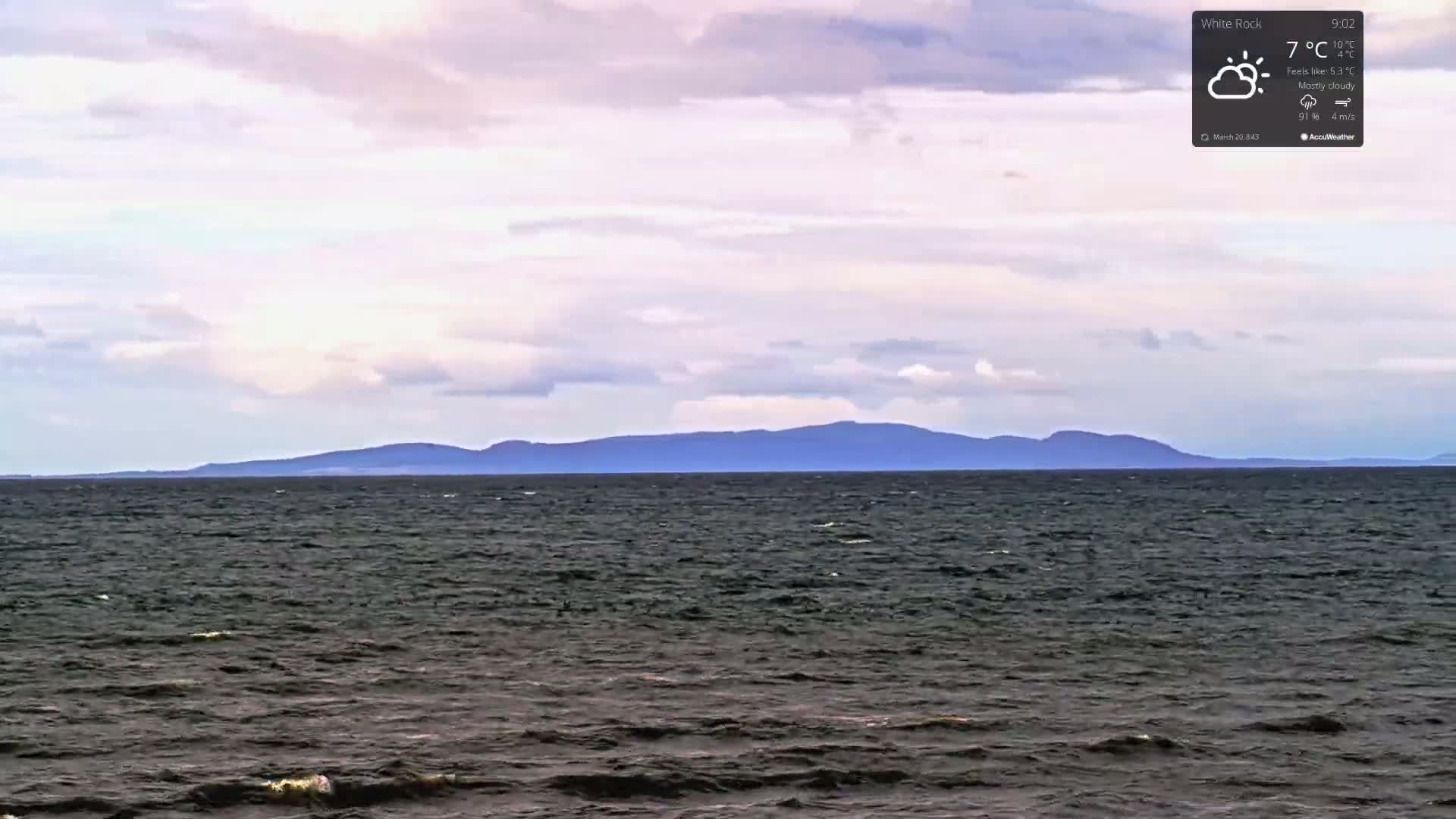 A mostly cloudy sky overlooks a dark, choppy ocean with a distant mountain range on the horizon.