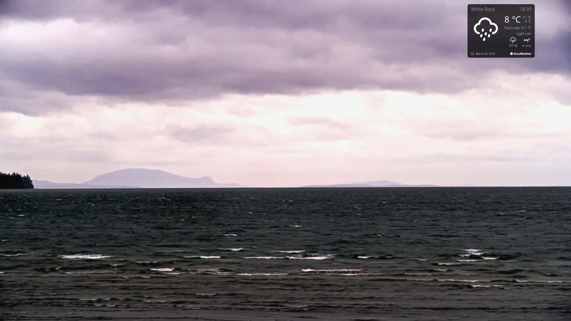 A dark, choppy sea stretches to a distant, low-lying landmass under a cloudy, overcast sky with light rain.