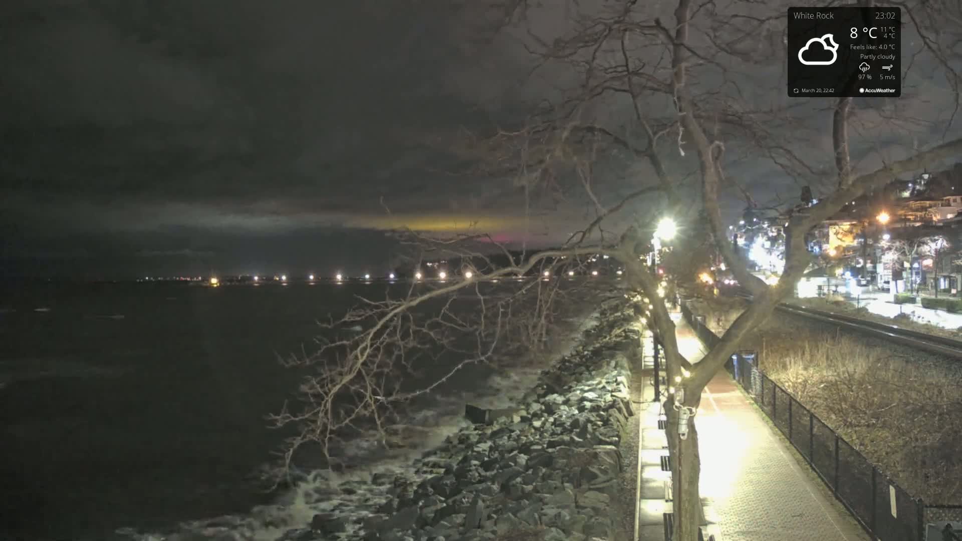 A nighttime, partly cloudy scene shows a rocky shoreline with waves lapping against it, a paved walkway lined with lights, and a town illuminated beyond a rail line.