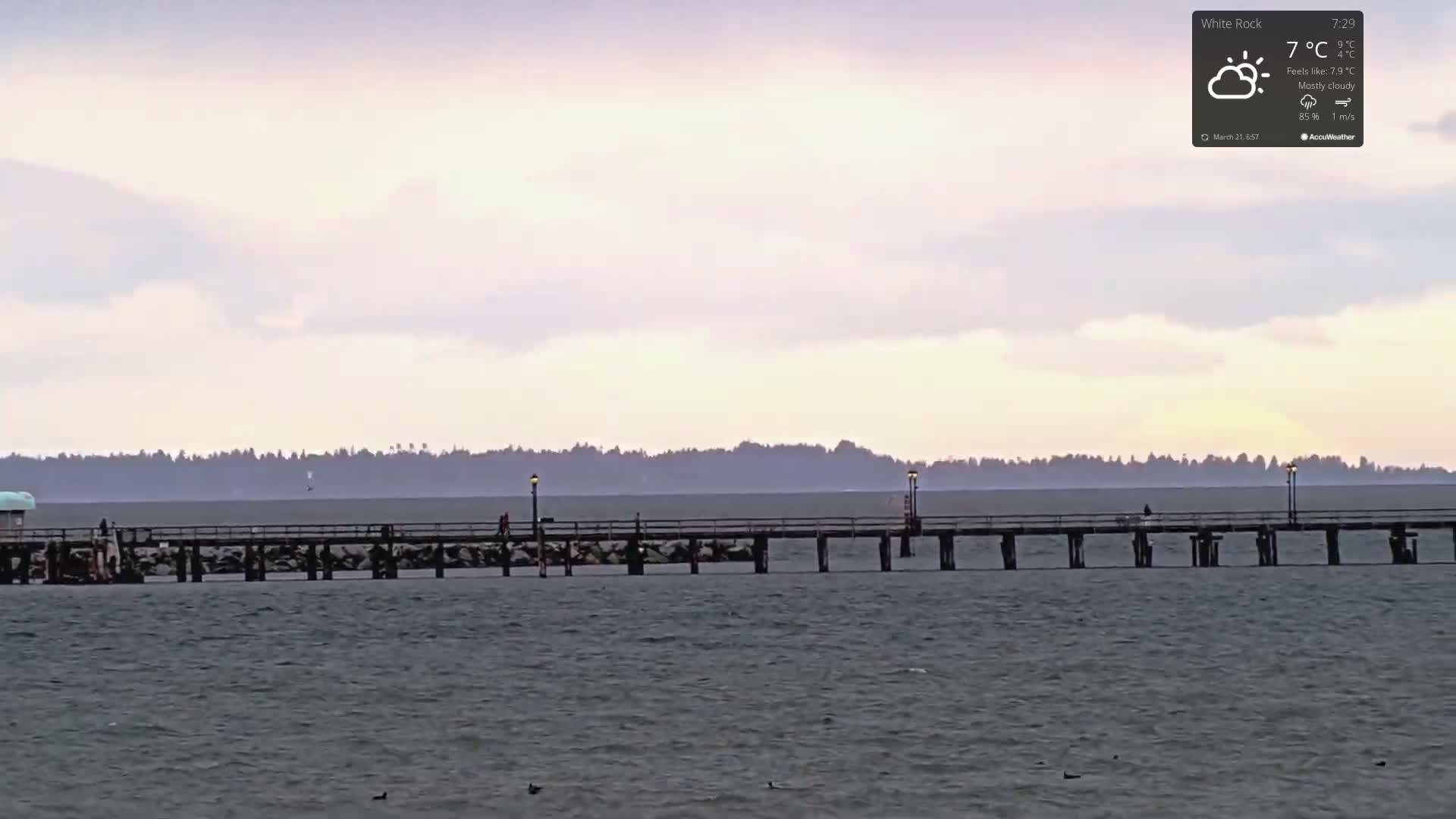 A long pier extends into a calm, grey body of water under a mostly cloudy, pale sky.