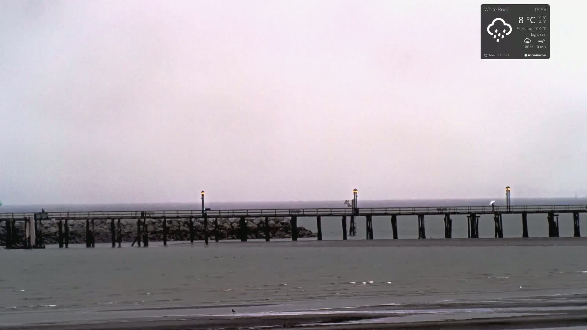 A long pier extends over calm, grey water under a light rain.