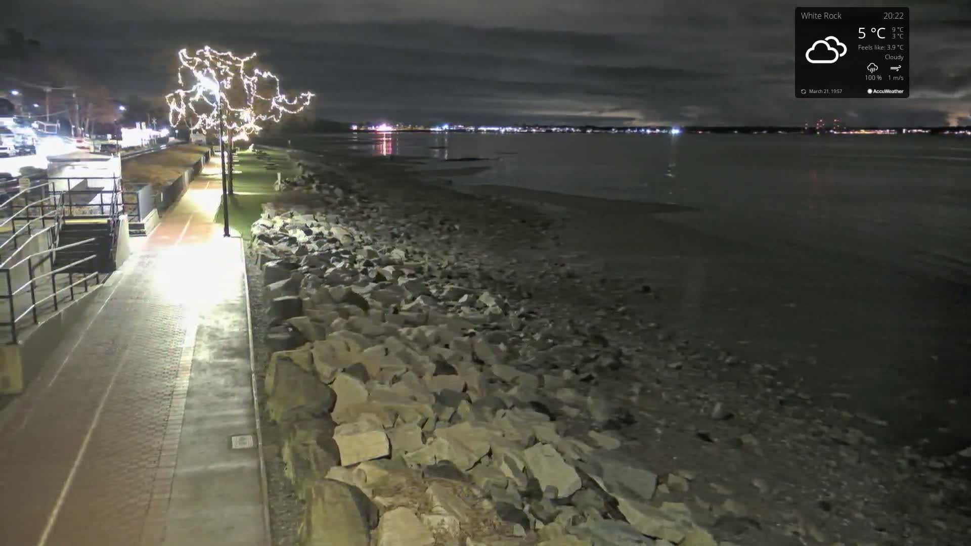 A nighttime, cloudy scene shows a paved walkway along a rocky shoreline with a body of water and distant city lights visible under a dark sky.