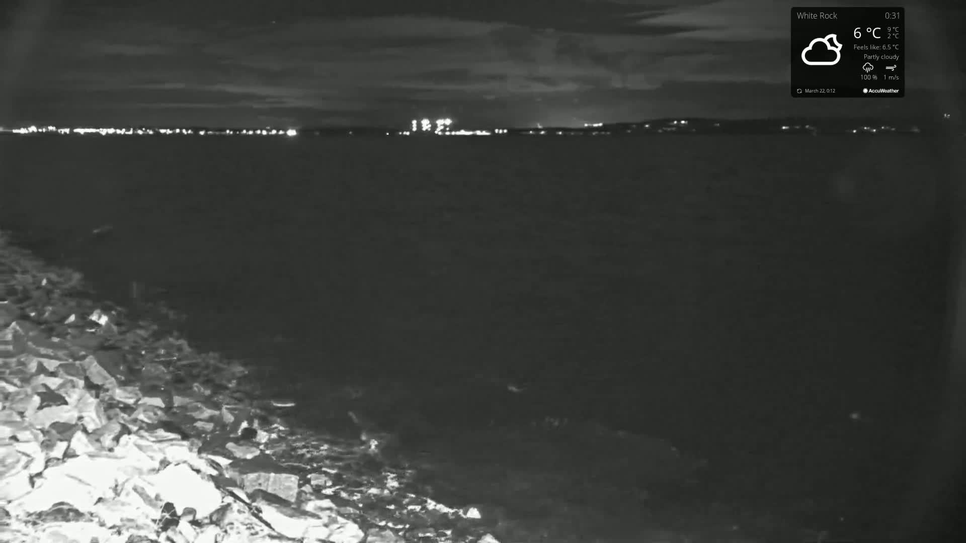 A nighttime, partly cloudy view of a dark body of water with distant city lights and a rocky foreground.