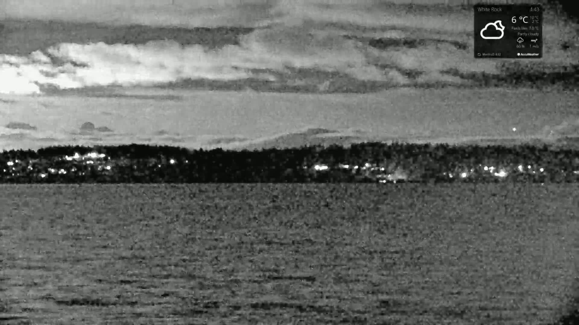A nighttime, partly cloudy view shows a body of water with a distant shoreline featuring a line of lights.