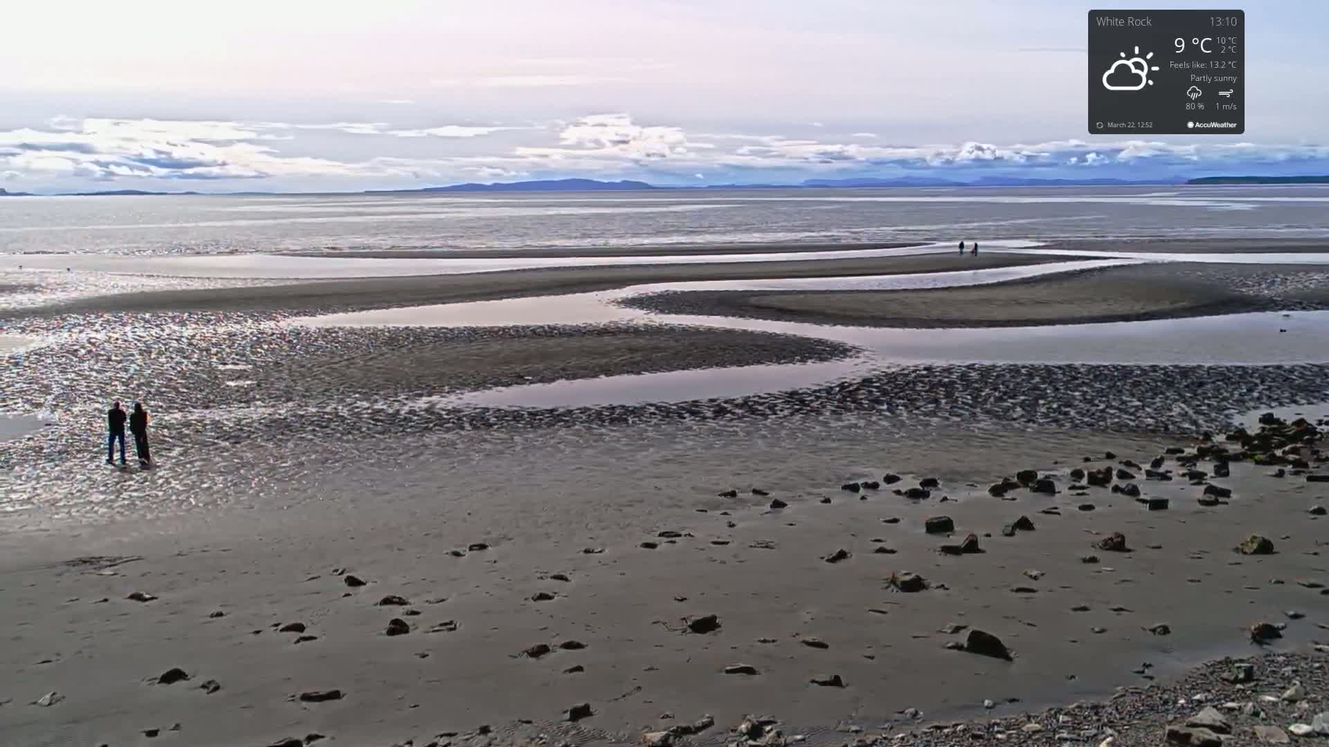 A wide, sandy beach with winding tidal pools stretches to a calm ocean under a partly sunny sky.