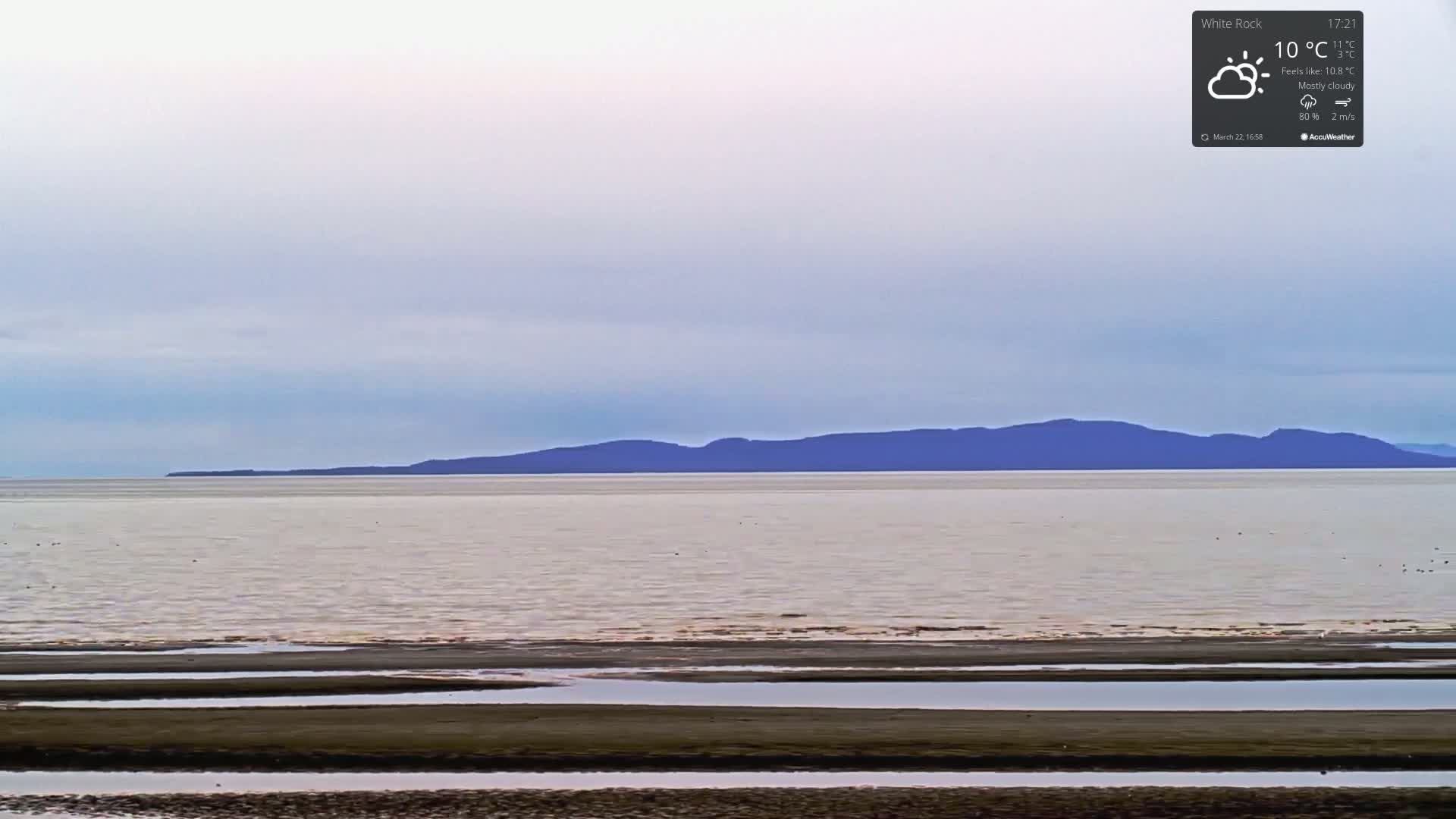A mostly cloudy sky overlooks a calm body of water with a dark mountain range in the distance, and a flat, muddy beach in the foreground.