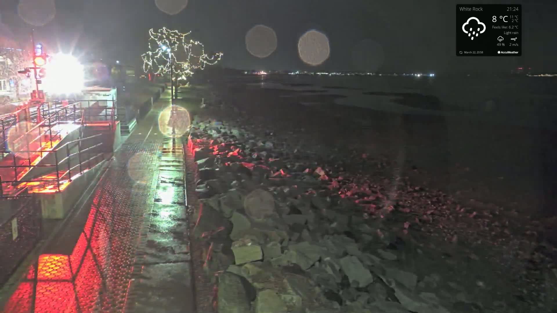 A wet, paved walkway lined with a metal railing and red lights at night leads to a rocky shoreline under light rain, with a decorated tree and distant lights visible.