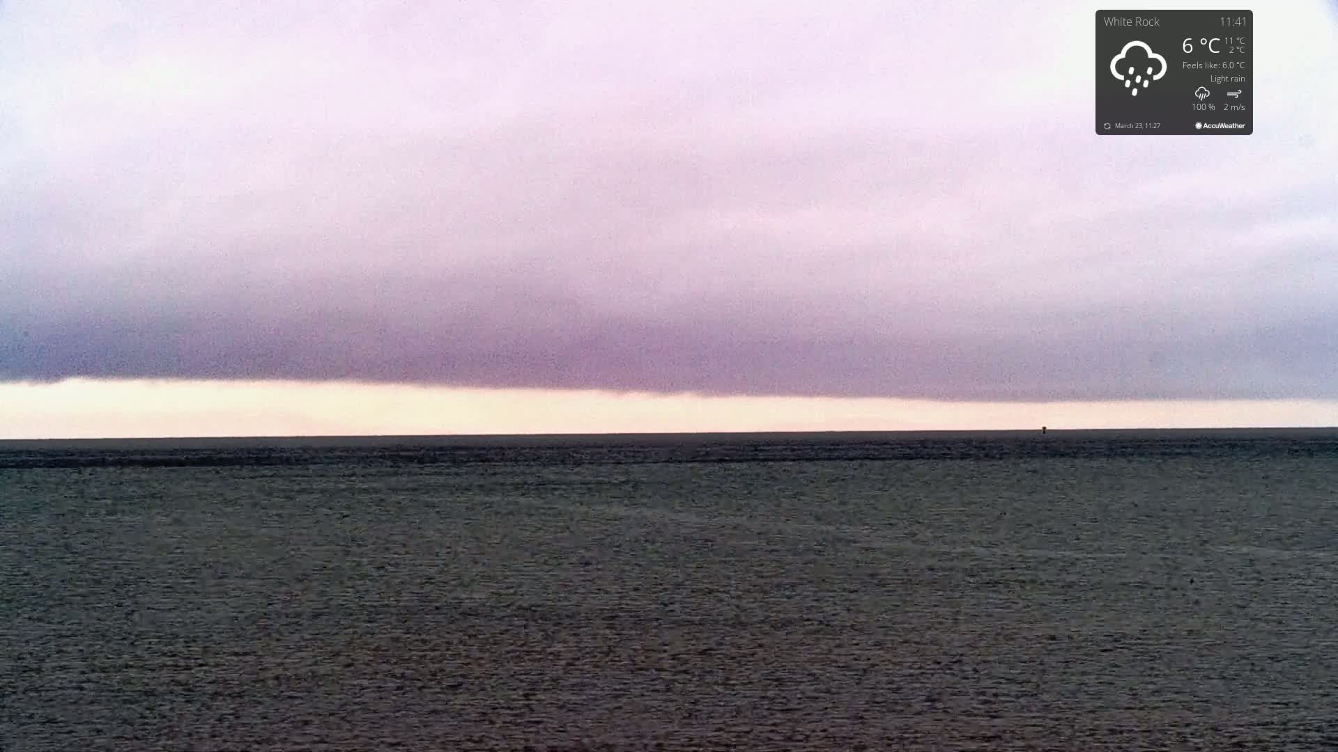 A dark, flat body of water stretches to the horizon under a mostly cloudy, purplish sky with light rain.