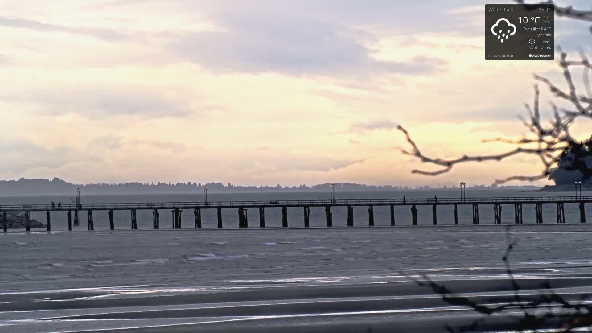 A long pier extends into a calm, gray body of water under a cloudy sky with light rain.