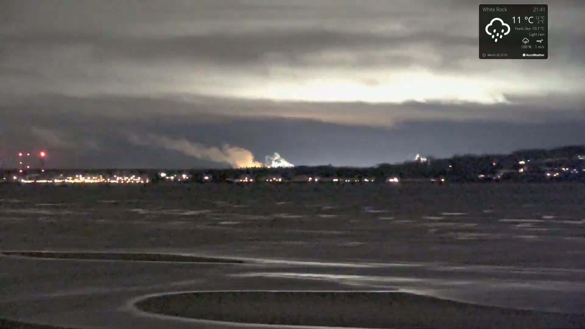 A nighttime, lightly raining scene shows a flat, dark expanse of land in the foreground, beyond which are distant lights of a town and industrial area under a cloudy sky.