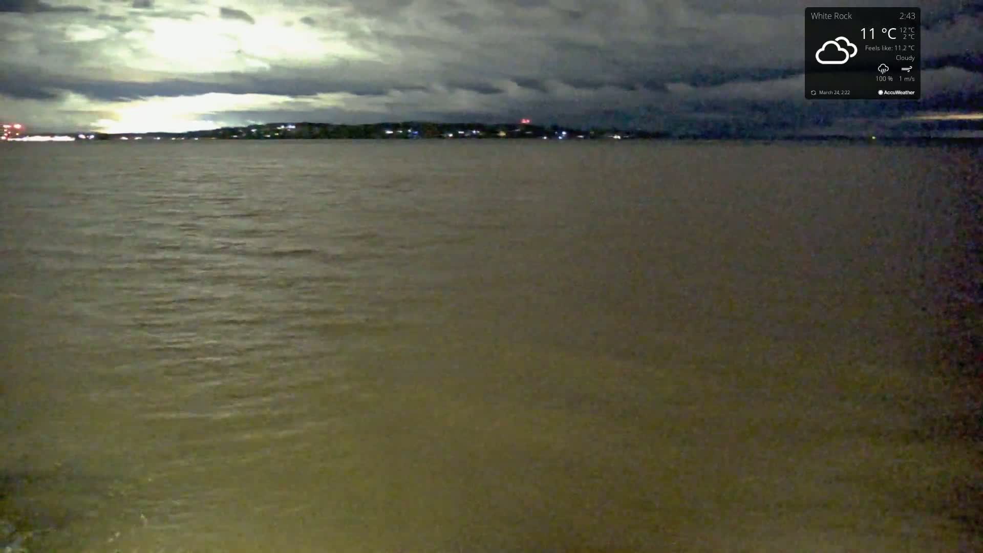 Under a cloudy night sky, a body of dark water stretches out to a dimly lit shoreline in the distance.