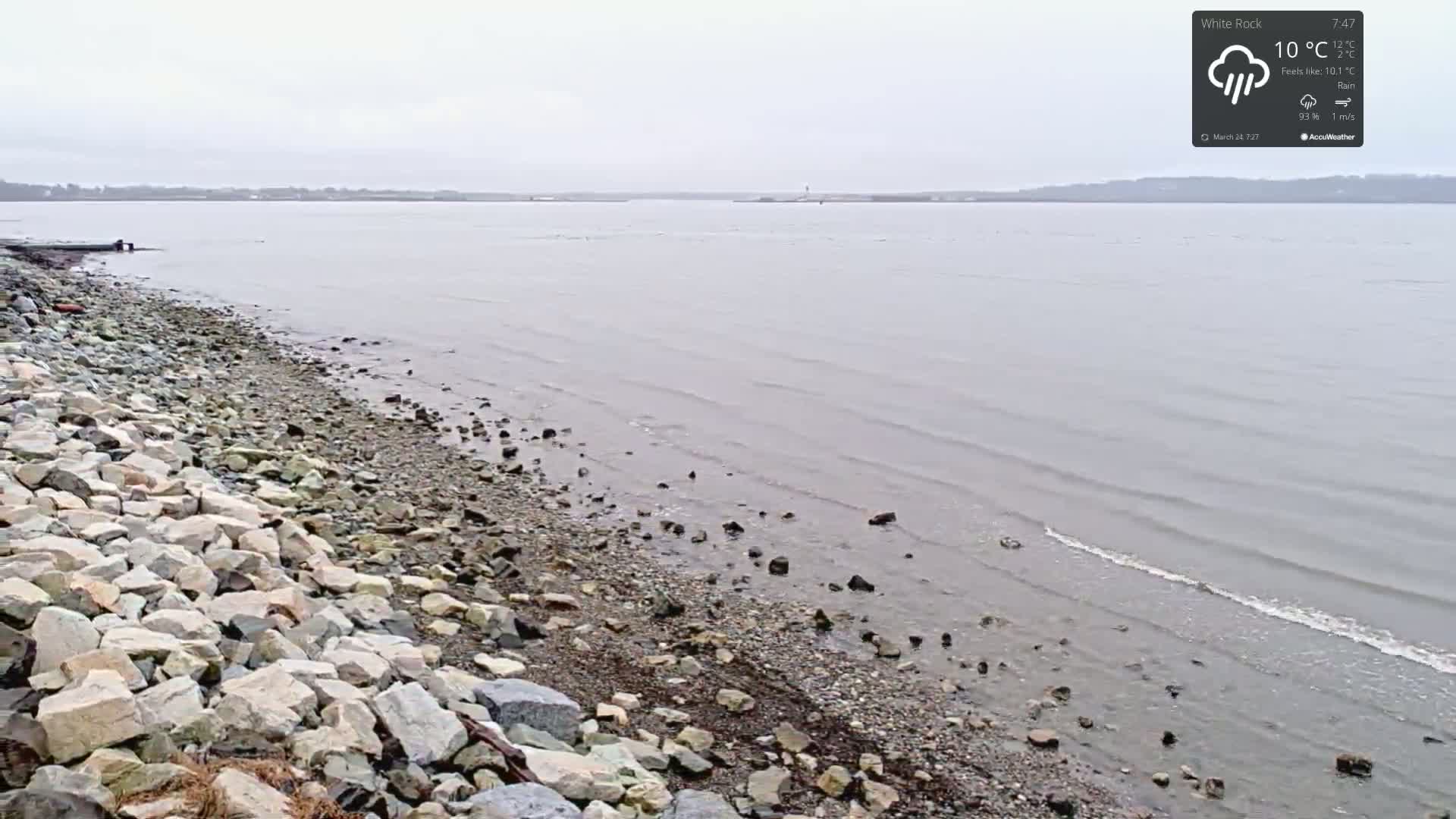A rocky shoreline meets a calm, grey body of water under a rainy, overcast sky.