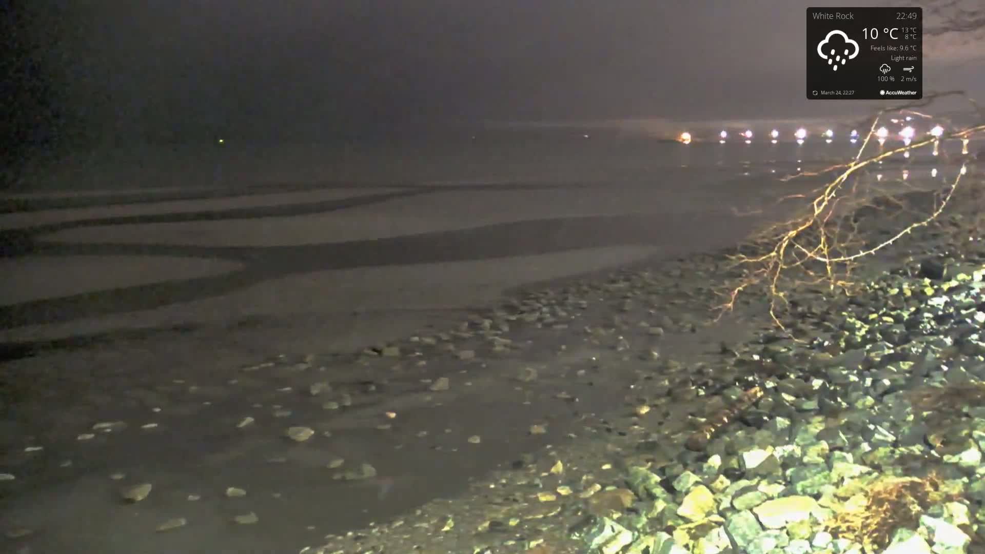 A dark, rocky shoreline is visible under a nighttime sky with light rain, and a series of lights are faintly seen in the distance across a dark body of water.