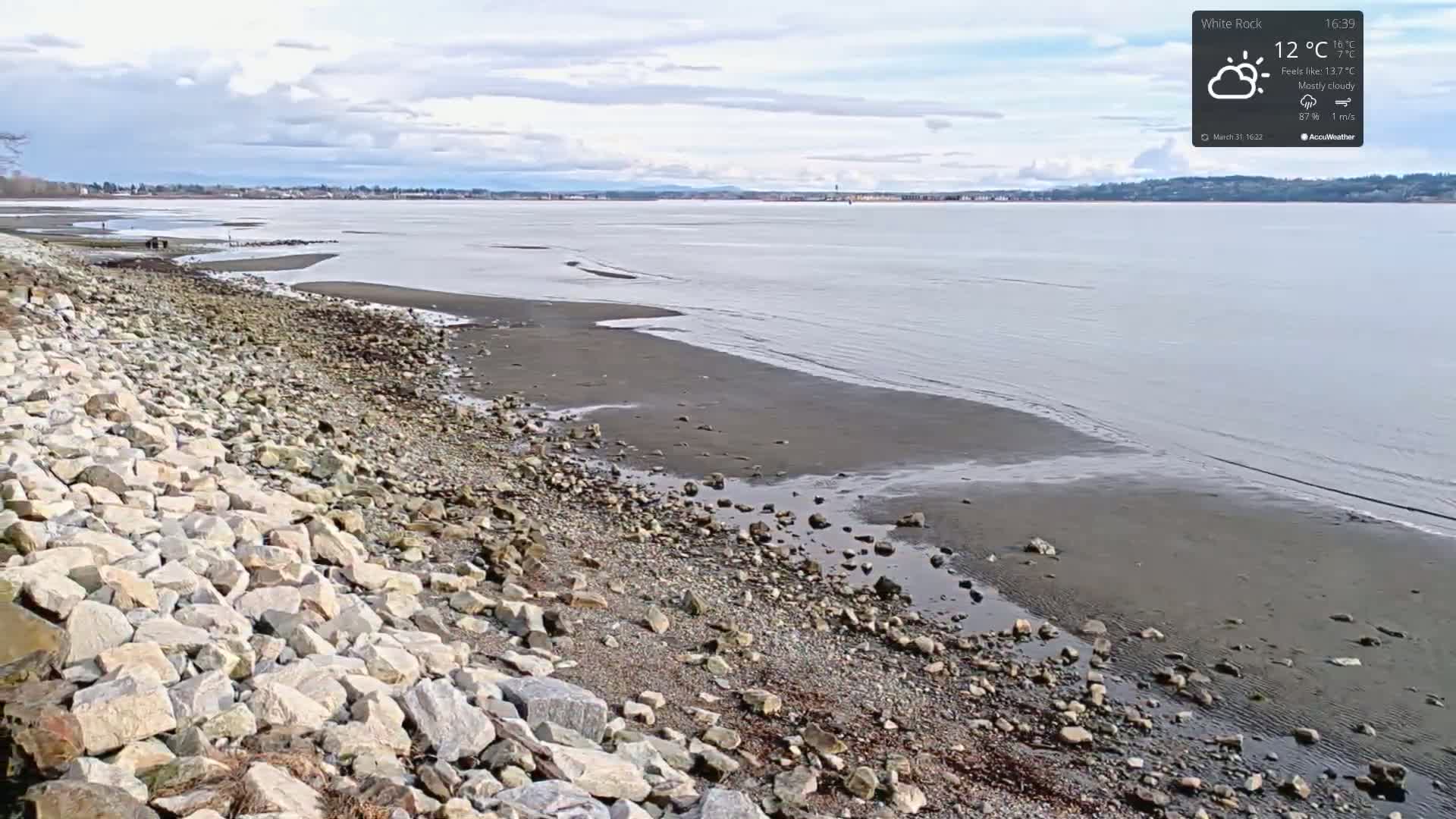 A rocky shoreline meets a calm, mostly cloudy body of water under a partly cloudy sky.