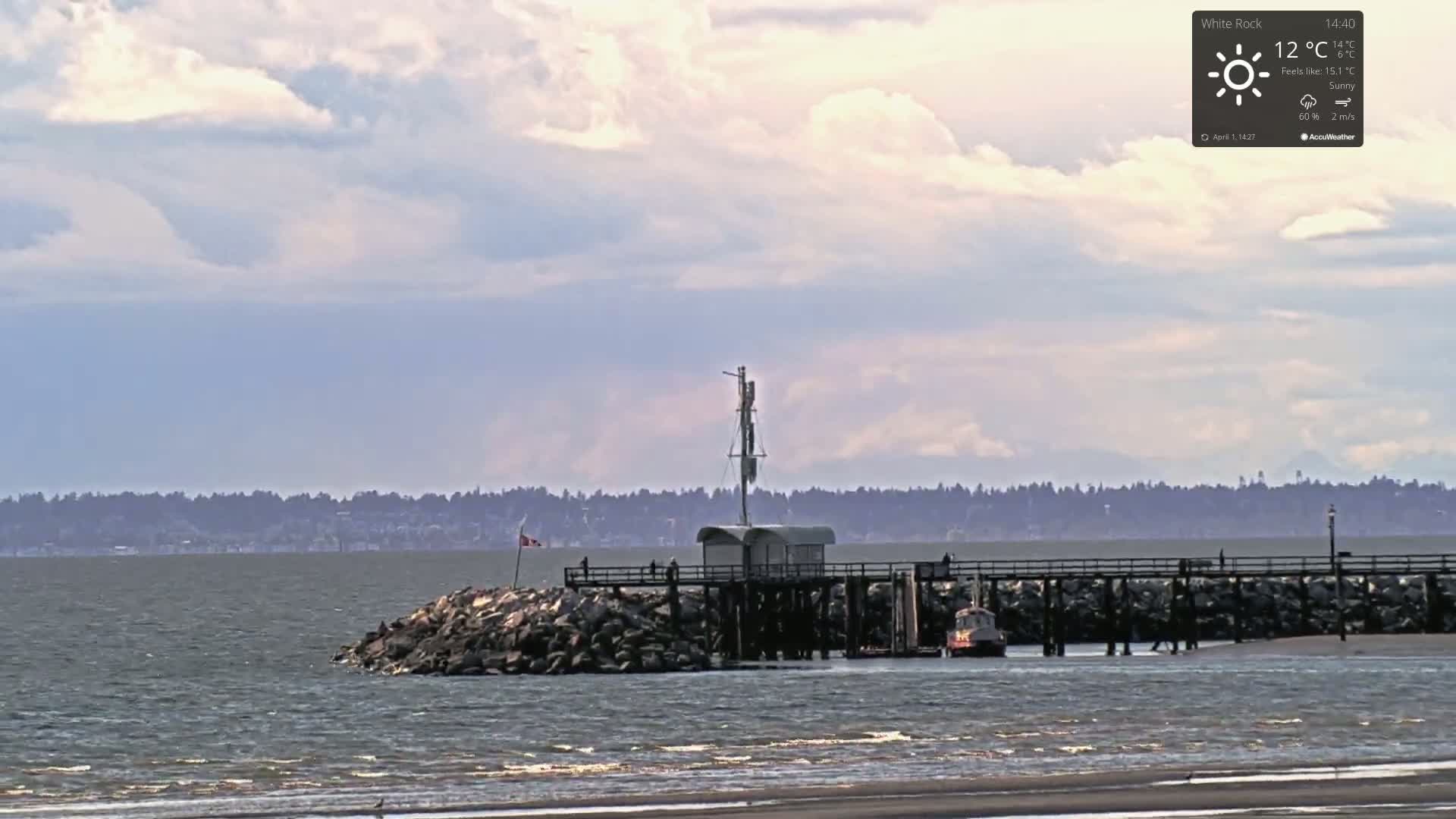 A pier extends into a calm body of water under a partly cloudy sky, with a distant tree line visible on the horizon.