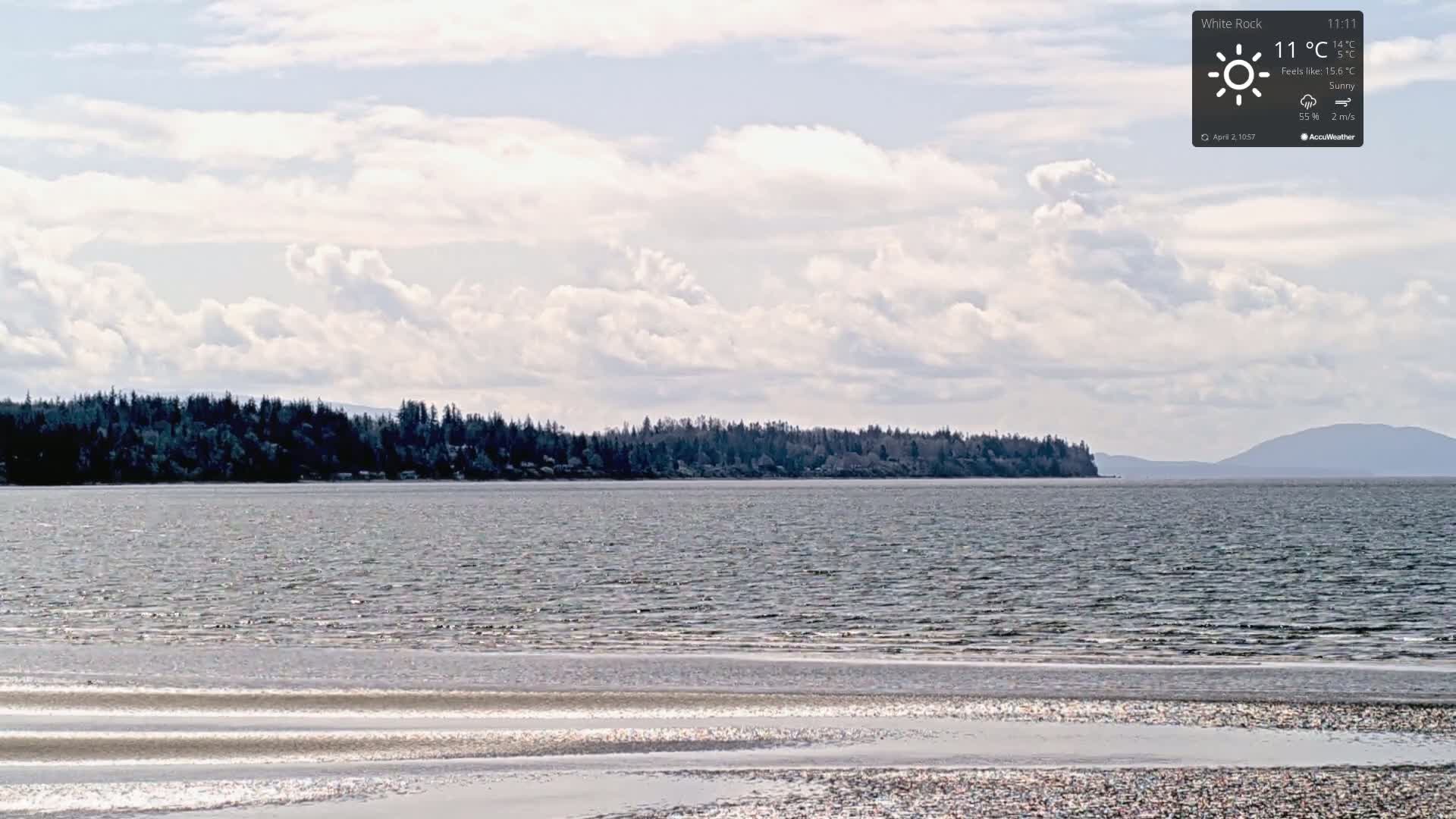 A partly cloudy sunny day shows a calm body of water with a shoreline of dark trees and distant mountains.