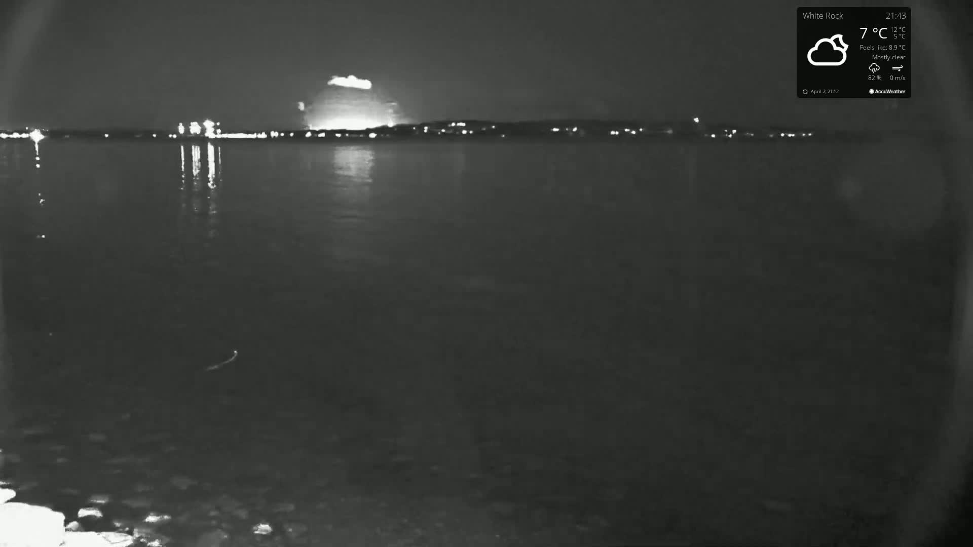 A nighttime view of a calm body of water reflecting the lights of a distant city under a mostly clear sky.