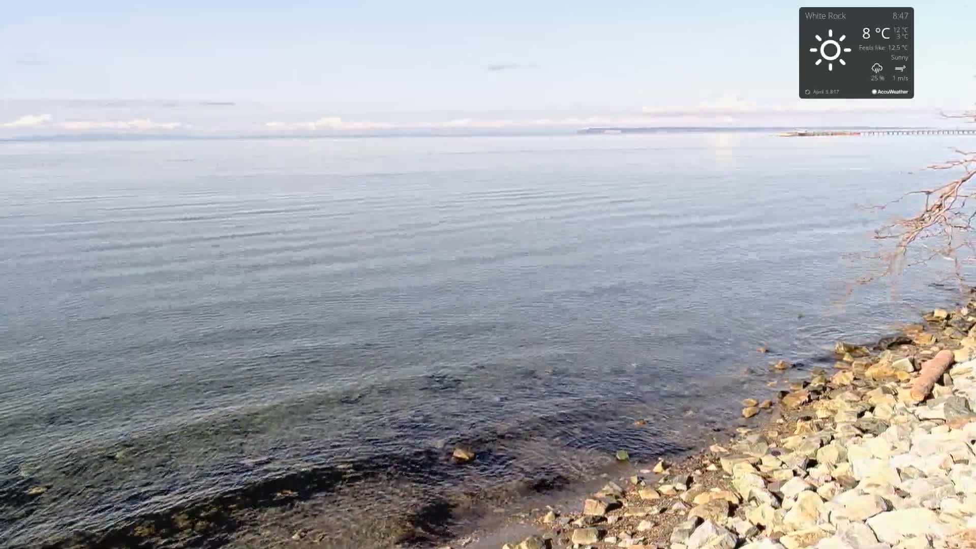 A rocky shoreline meets a calm, sunny ocean under a mostly clear sky.