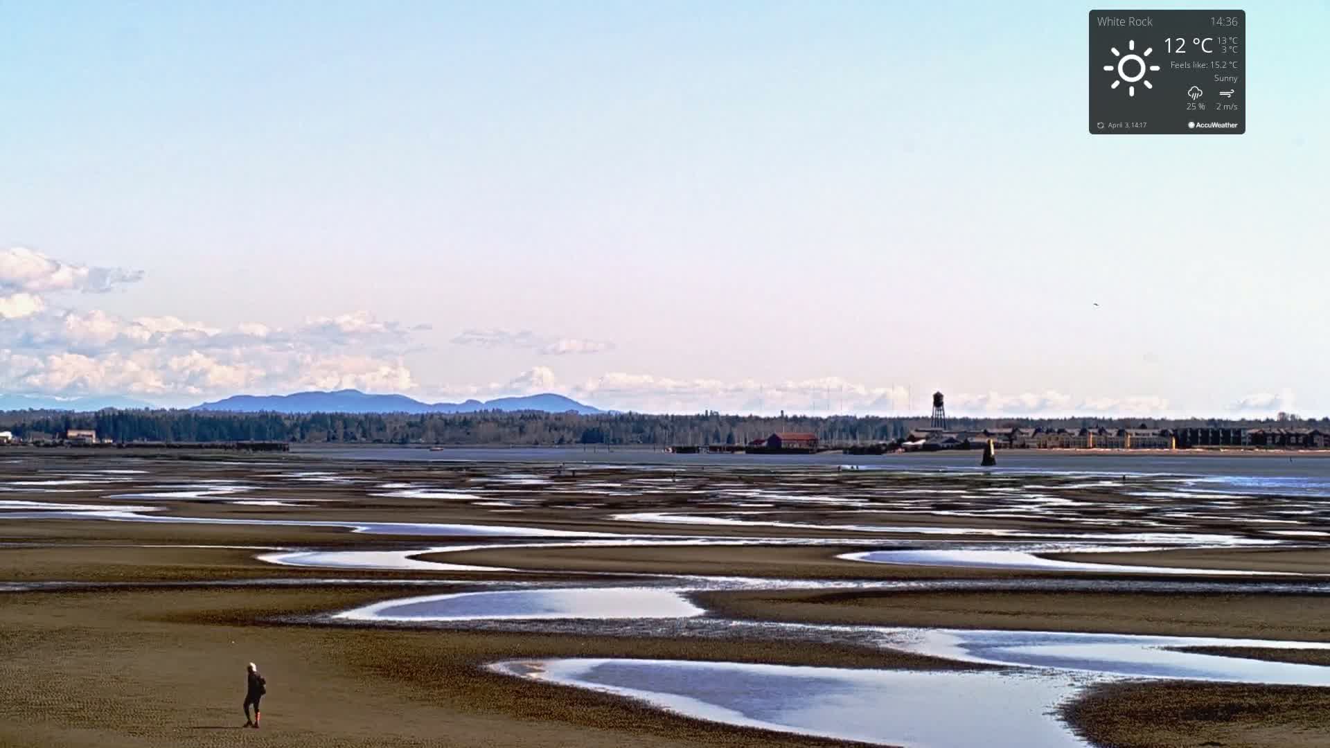 A person stands on a wide, flat, sandy expanse patterned with numerous shallow pools of water under a mostly clear, sunny sky, with distant mountains visible on the horizon.