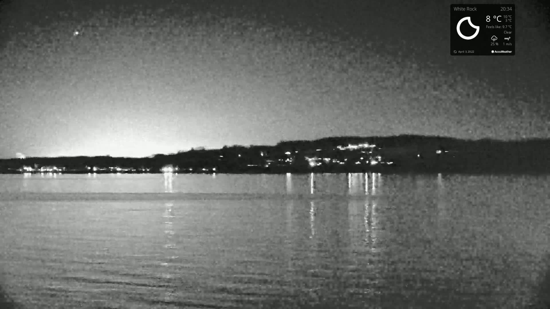 A nighttime, clear view of a calm body of water reflecting the lights of a distant town under a dark sky.