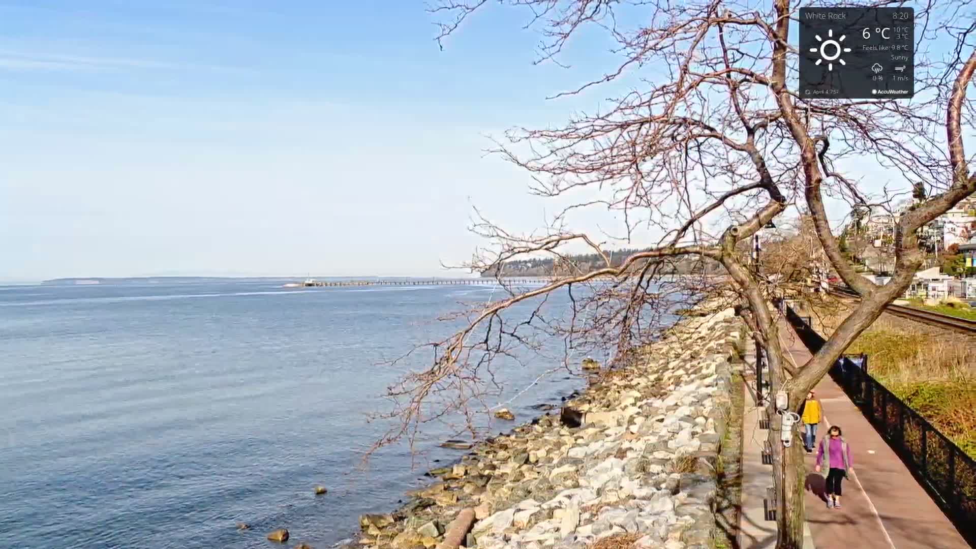 Two people walk along a seaside path on a sunny day, with a rocky shoreline and calm water extending to a distant landmass.