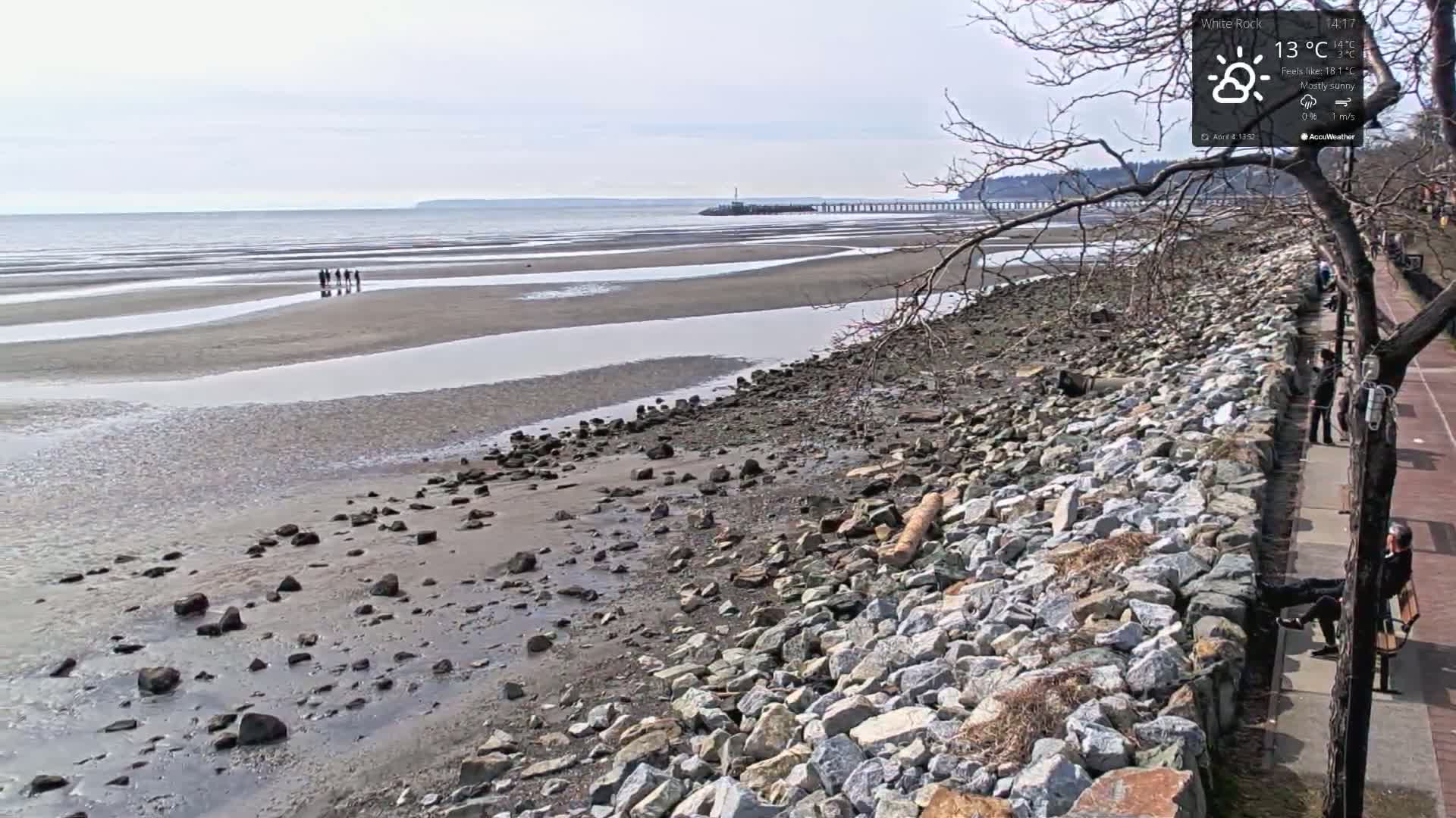 A mostly sunny day reveals a rocky shoreline with a few people walking on a wide, wet, sandy beach, a distant pier, and calm water under a pale sky.