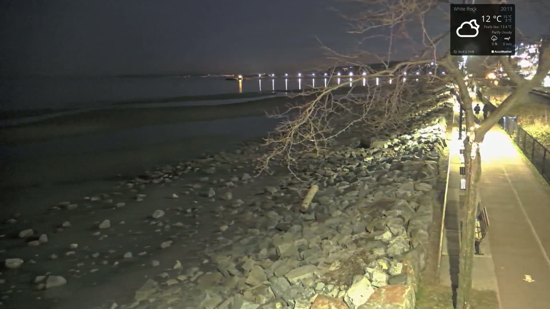 A rocky shoreline at night under partly cloudy skies is illuminated by streetlights lining a paved path beside it, with a calm body of water visible in the distance.