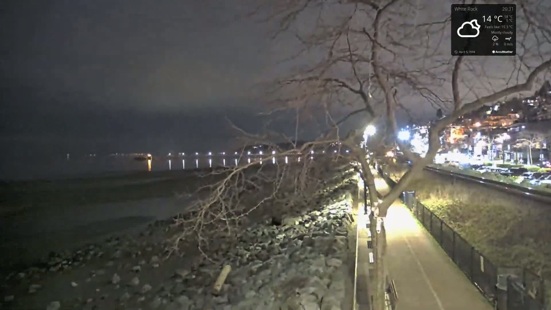 A nighttime view of a paved waterfront path next to a rocky shoreline, under a mostly cloudy sky, with a calm body of water and distant lights across it.