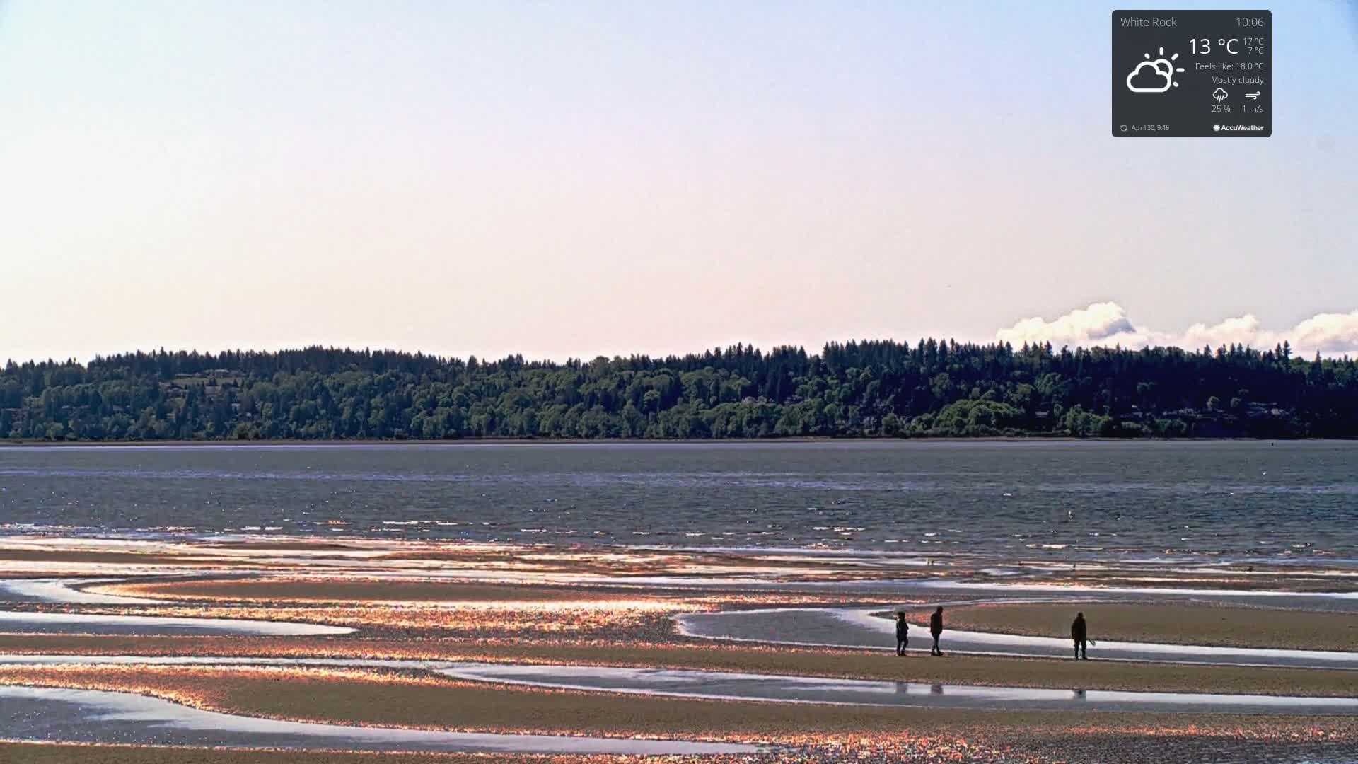 A mostly cloudy sky overlooks a calm body of water with a sandy shoreline showing tidal pools; three people walk on the exposed sand.