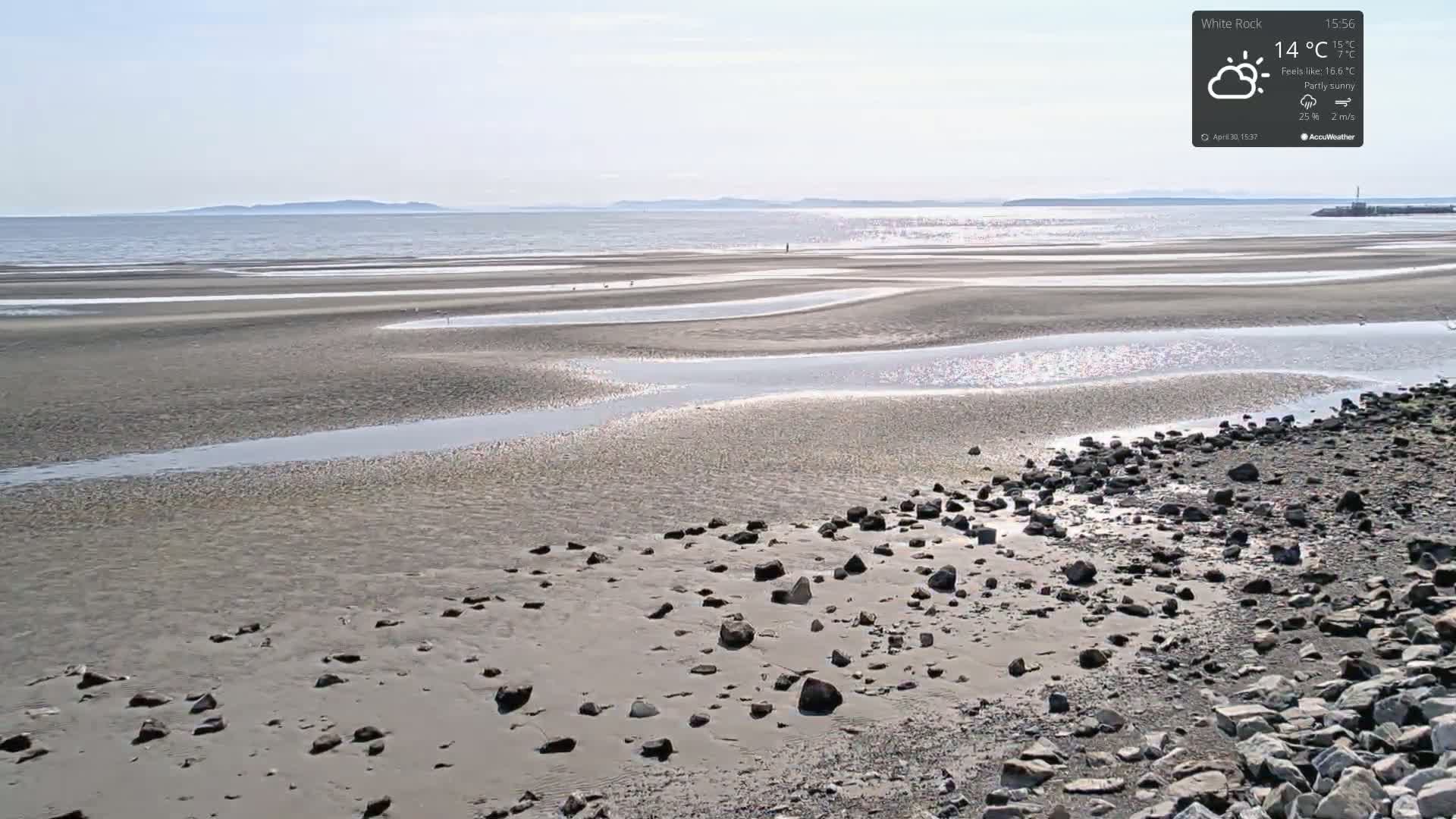 A partly sunny beach with a calm sea and winding tidal channels shows scattered rocks in the foreground.