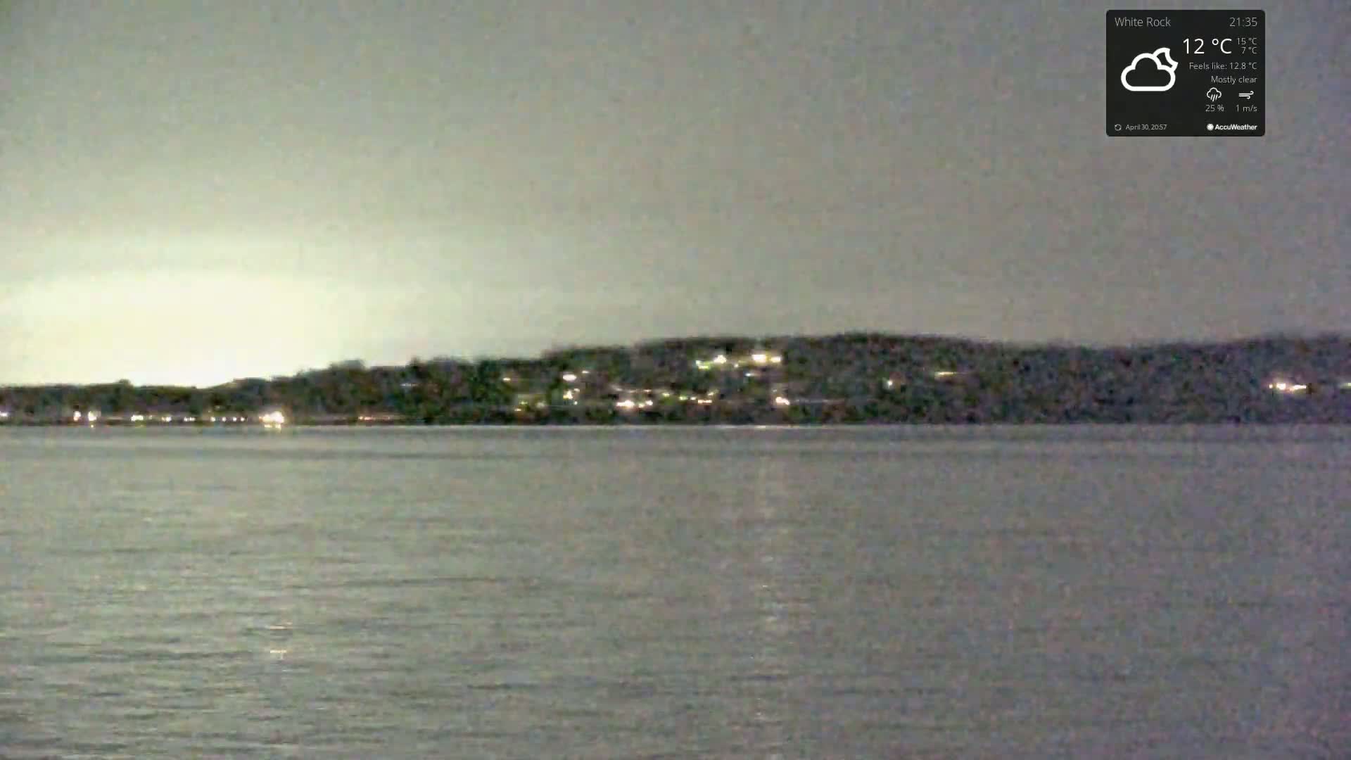 A mostly clear night shows a dark, hilly shoreline dotted with lights across a calm body of water.