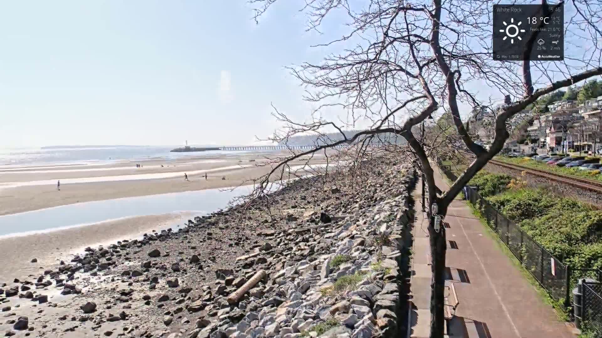 A rocky shoreline with a paved path and a few people on a sunny beach next to a calm body of water, under a mostly clear sky.