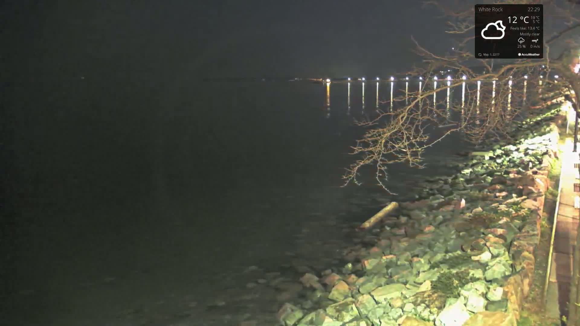 A dark, mostly clear night scene shows a rocky shoreline next to a body of water with lights faintly visible in the distance.