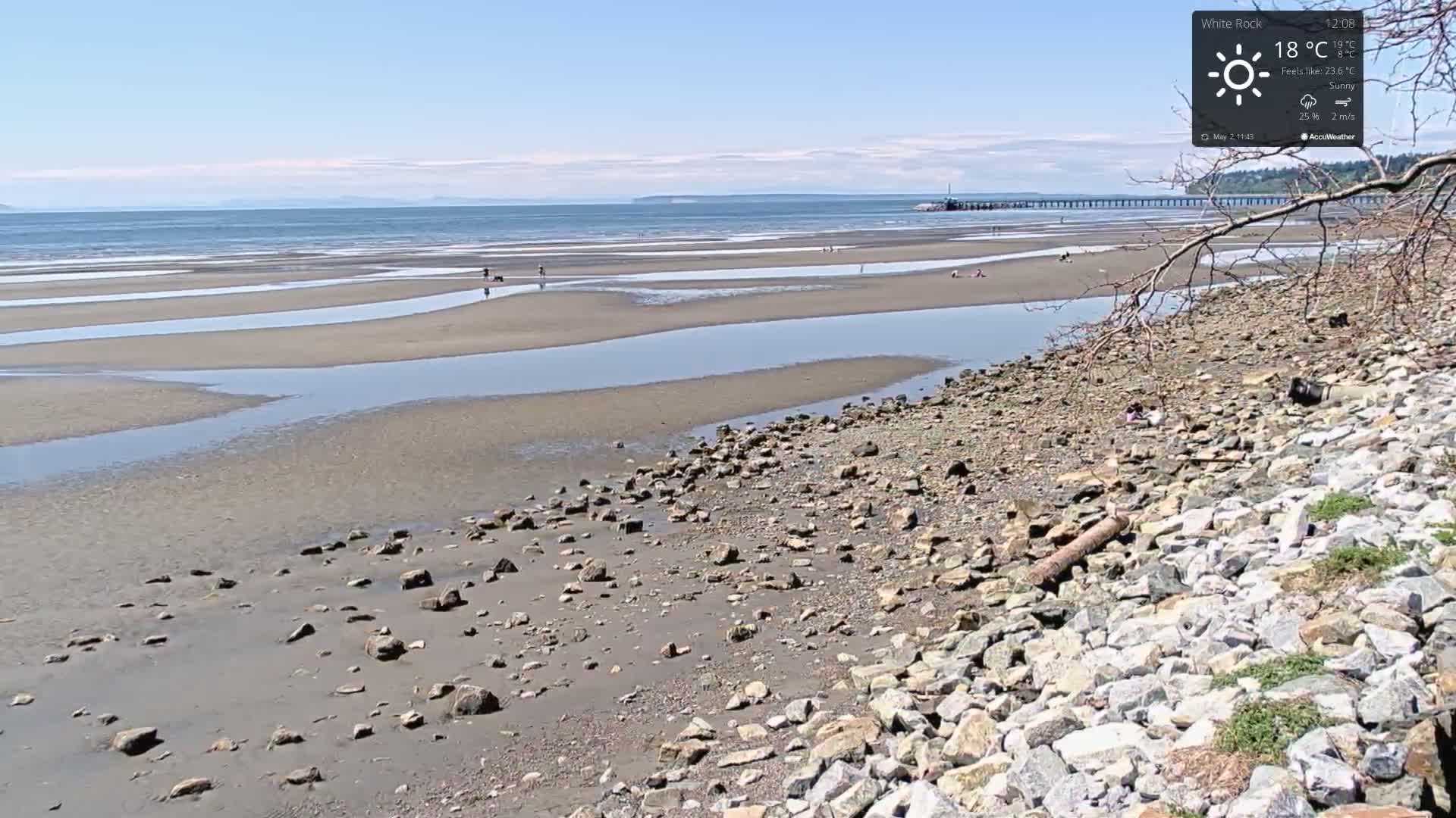A rocky shoreline with a sandy, tidal beach stretches toward a calm ocean under a sunny sky.